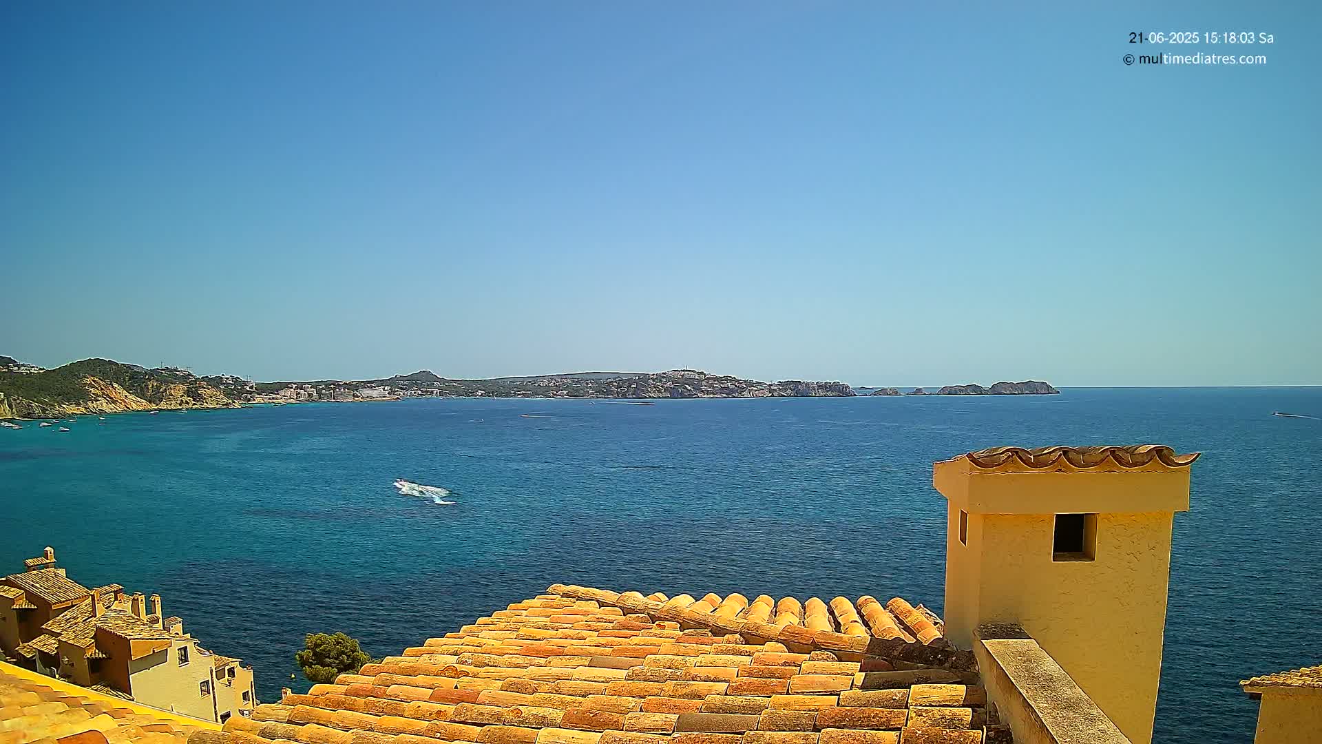 A sunny, clear day shows a coastal scene with a terracotta roof and a small building overlooking a calm blue sea, with land visible in the distance.