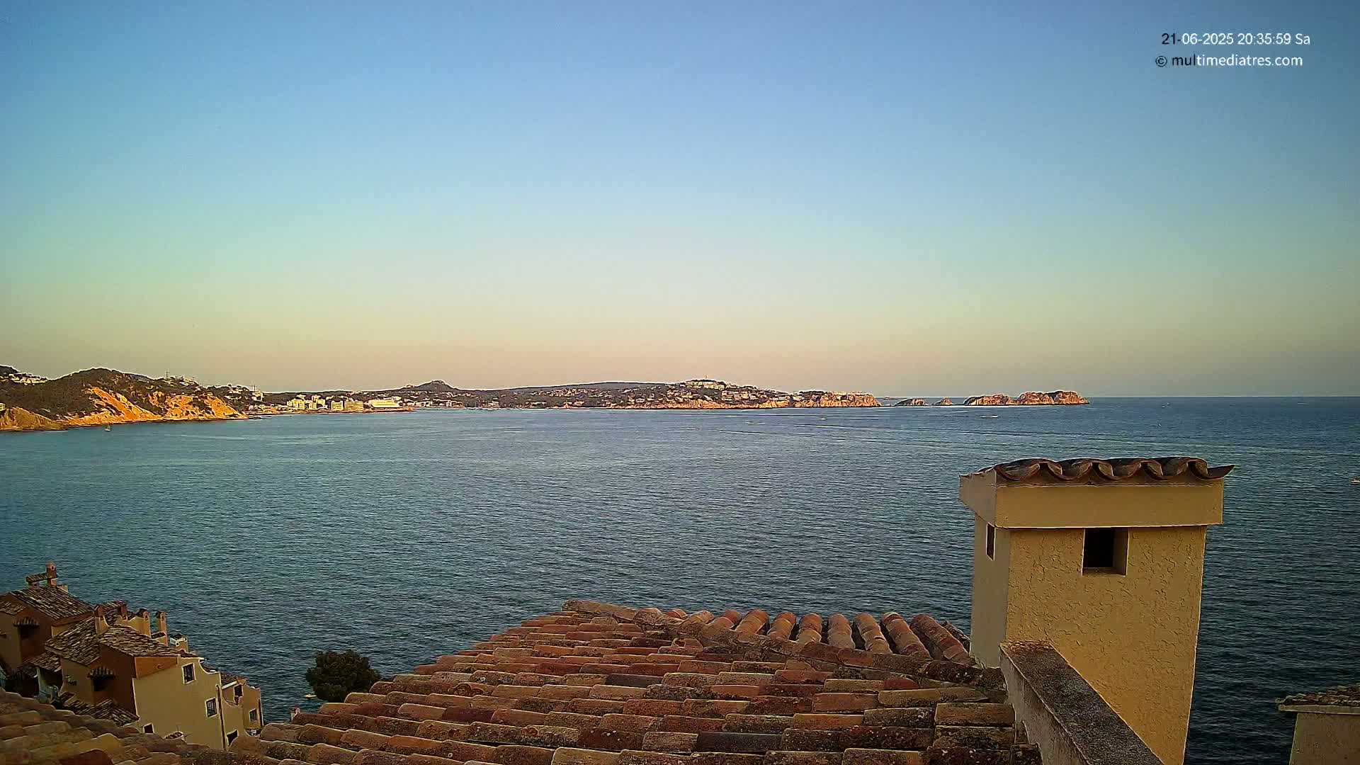 A calm sea with a distant coastline under a clear, twilight sky is viewed from a rooftop with terracotta tiles.