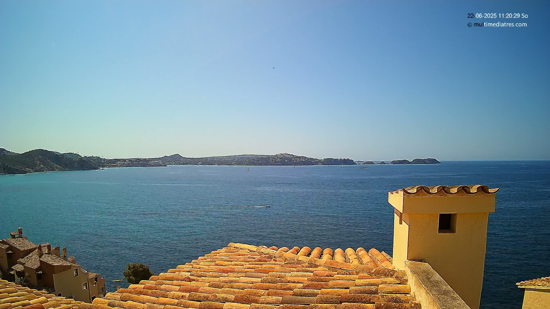 A terracotta roof overlooks a calm blue sea with distant land under a clear blue sky.