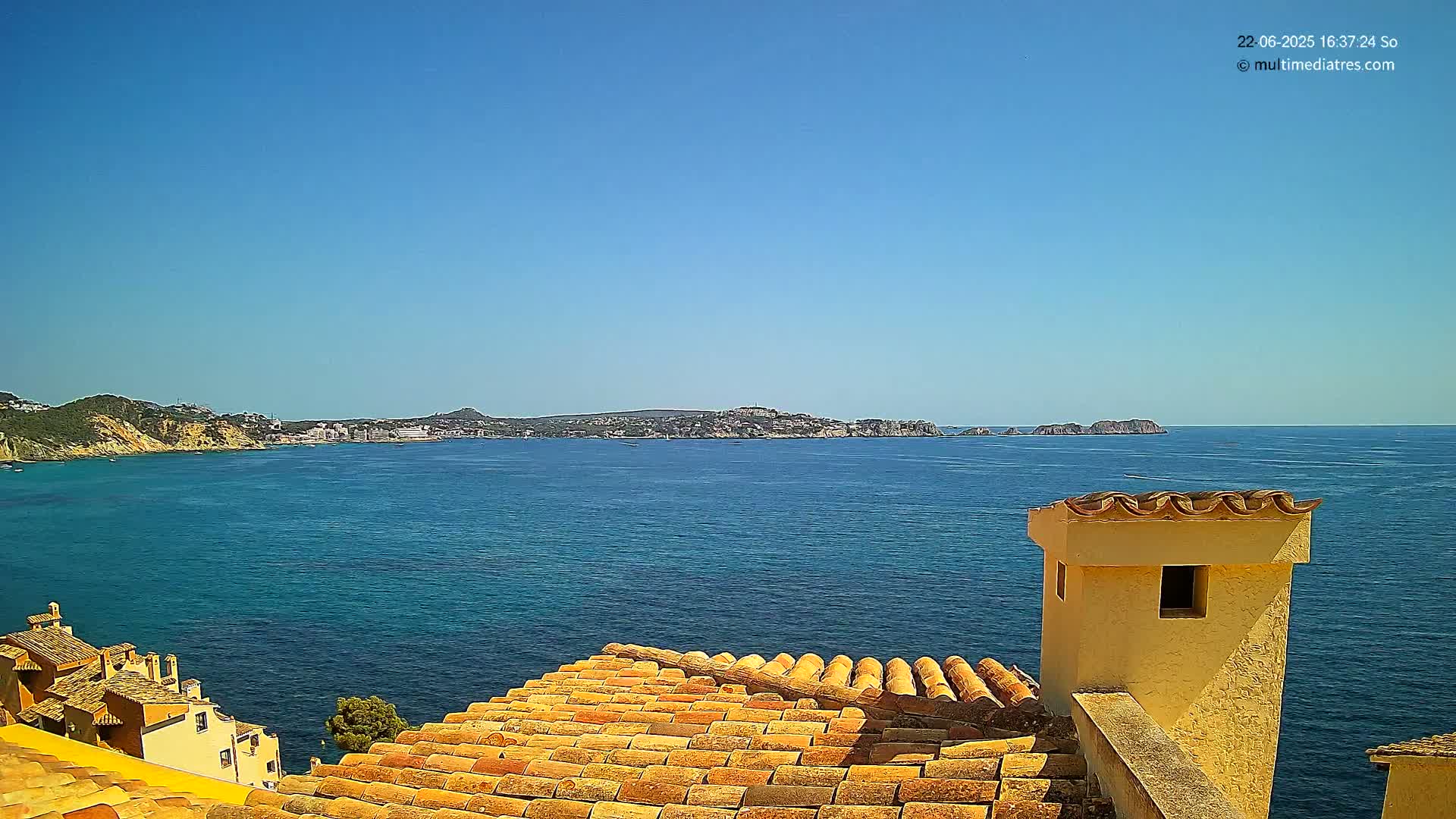 A terracotta roof overlooks a calm blue sea and distant coastline under a clear blue sky.