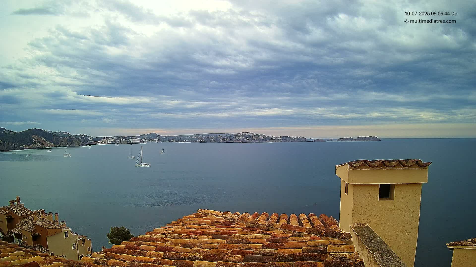 A terracotta roof overlooks a calm sea with several sailboats under a mostly cloudy sky.