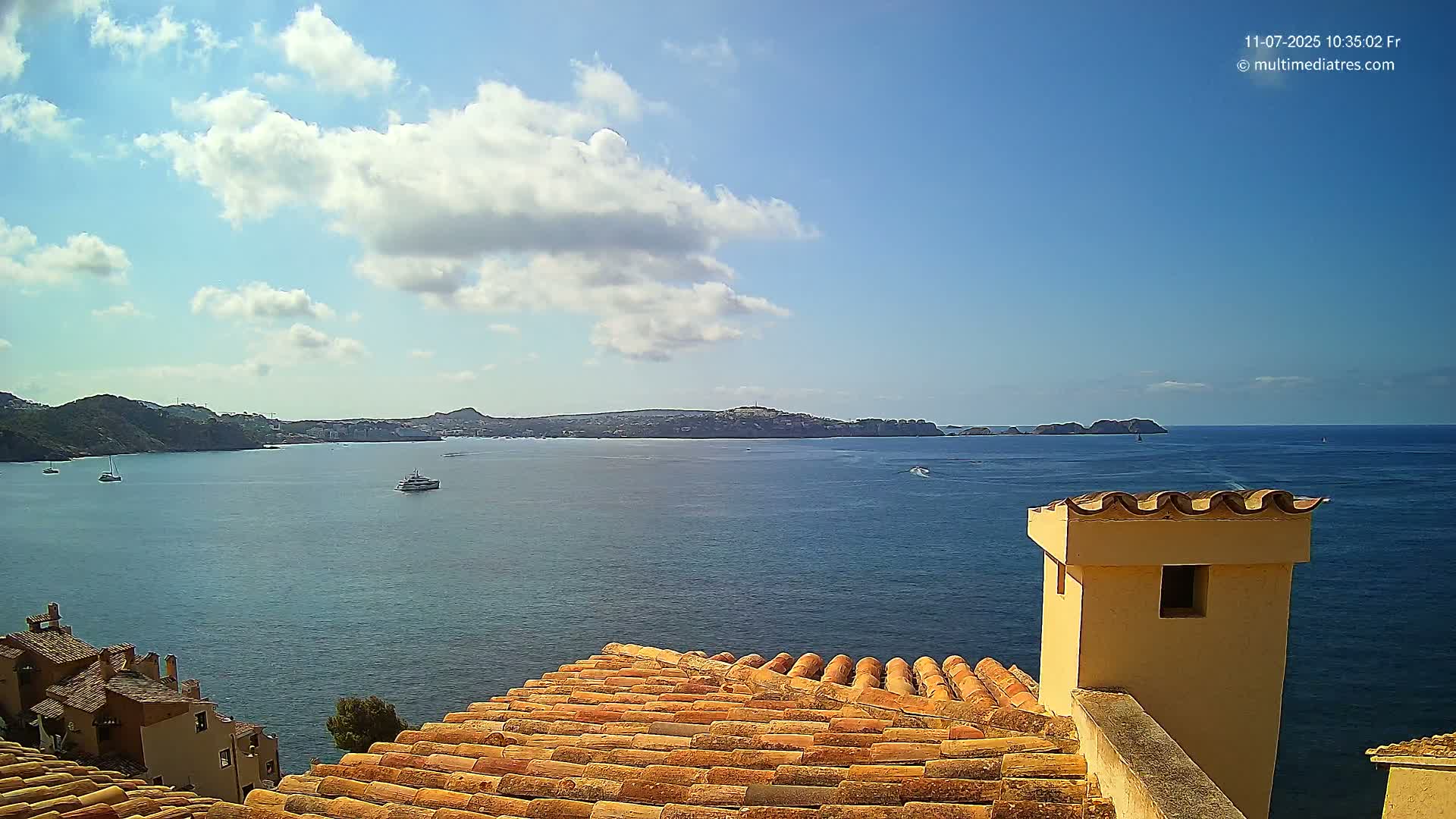 A partly cloudy sky overlooks a calm bay with several boats, viewed from a terracotta roof with a short, pale yellow chimney.