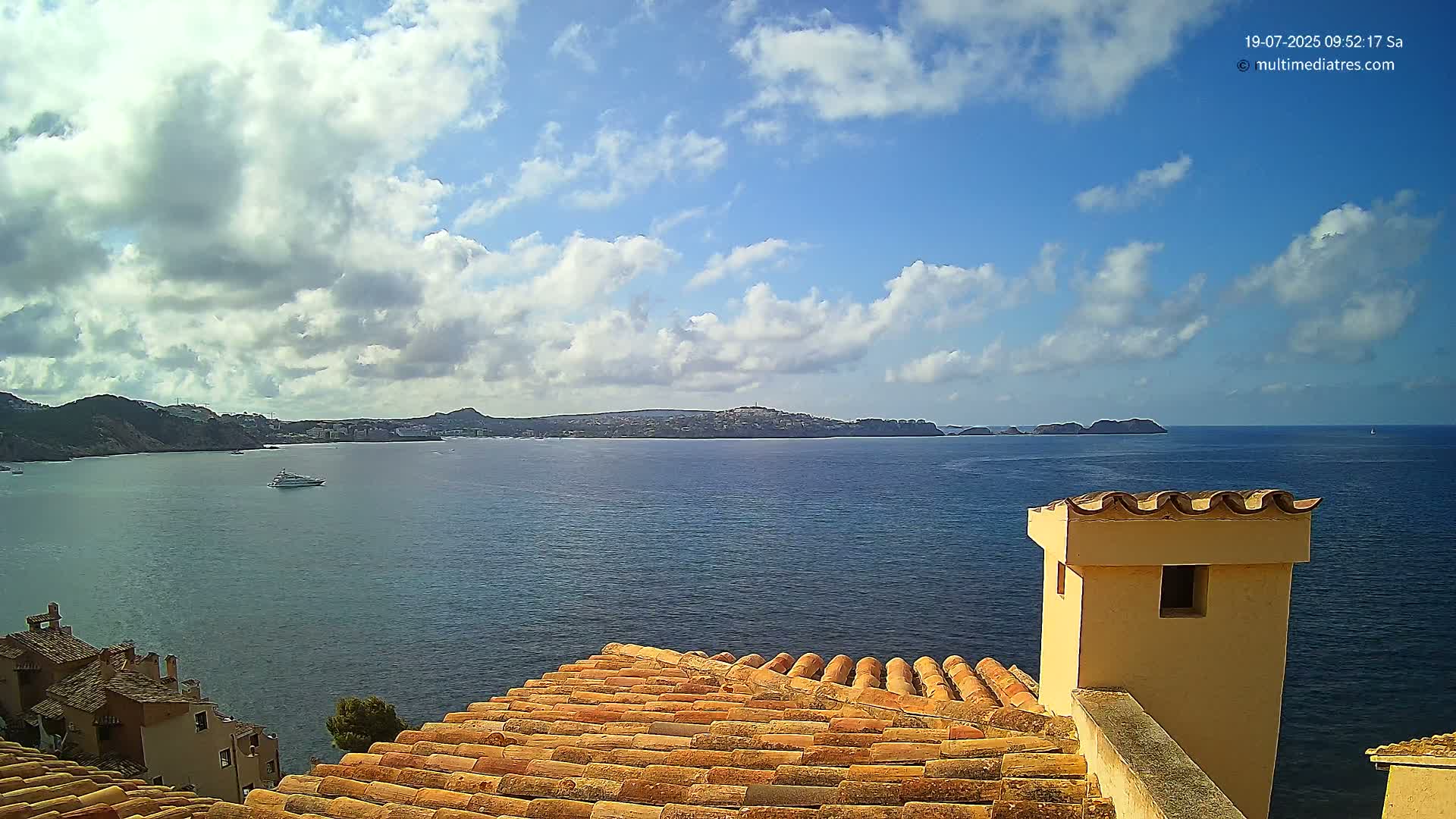 A partly cloudy blue sky overlooks a calm ocean, a distant coastline, and a terracotta roof with a small chimney.