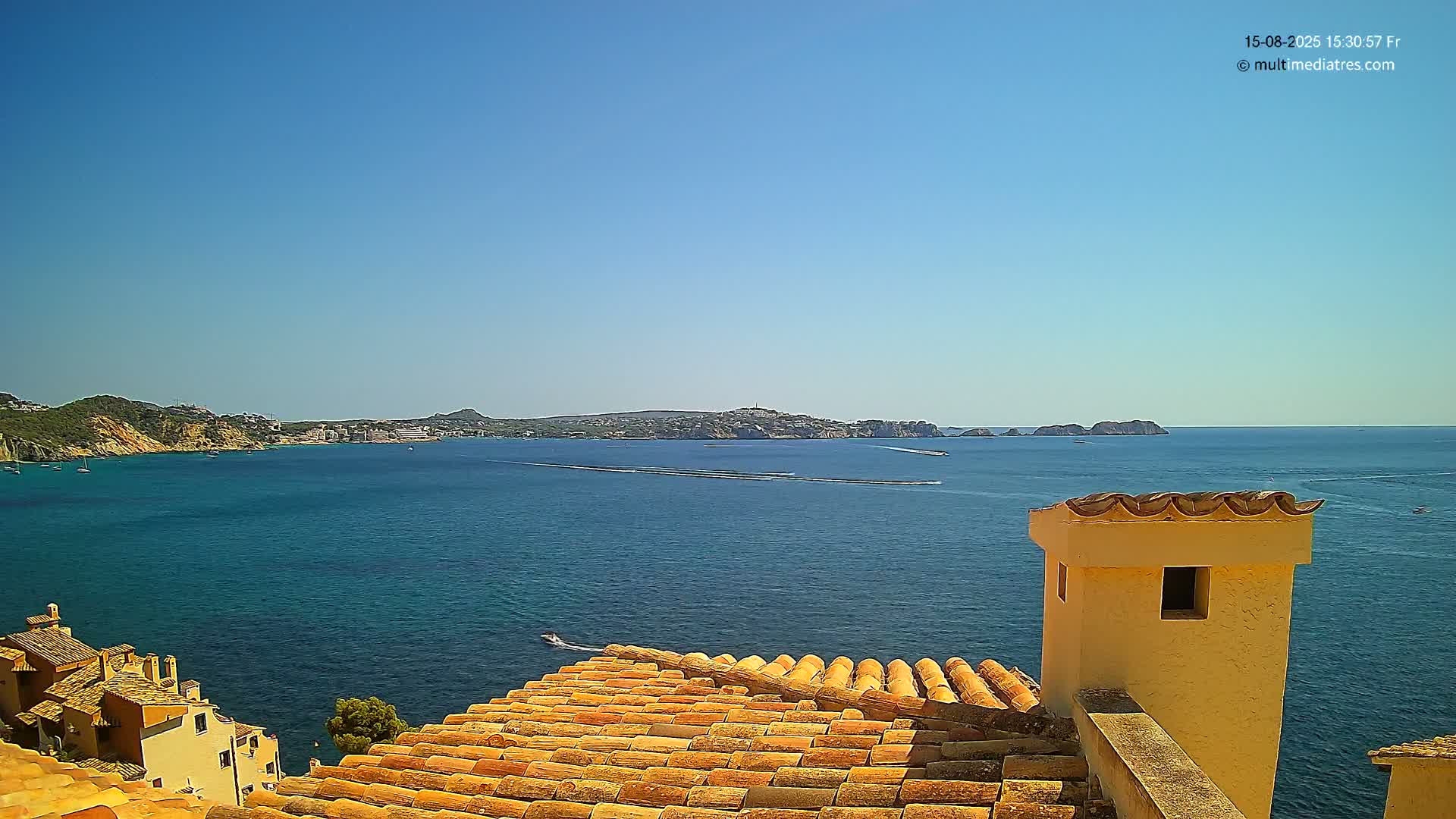 A view from a terracotta roof shows a calm, blue sea with distant land and islands under a clear blue sky.