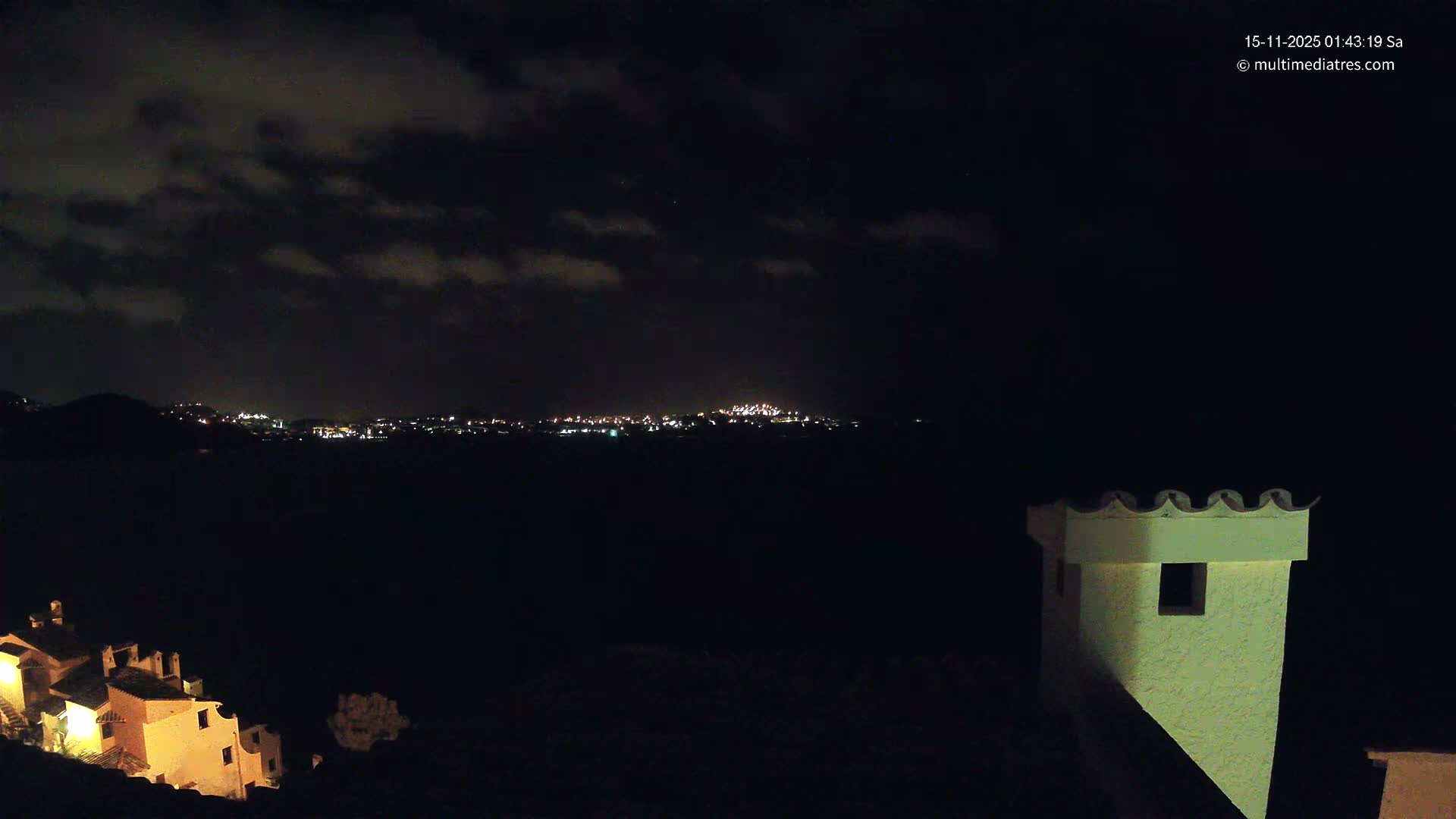 The image displays a cloudy night view of a distant illuminated city skyline across a dark expanse, with several warmly lit buildings on the lower left and a prominent, light-colored turret structure on the lower right.