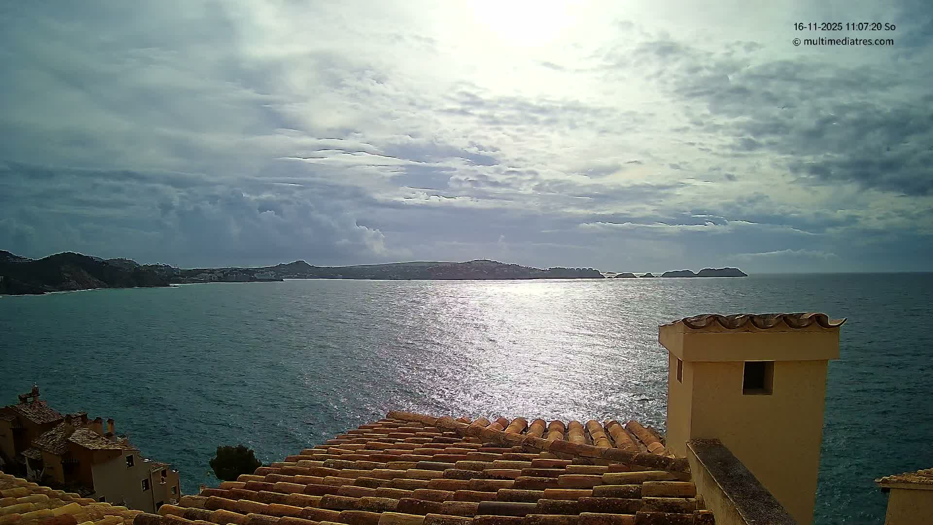 Terracotta rooftops and a chimney frame a sparkling blue sea leading to a distant hilly coastline and small islands, all under a dramatic, partly cloudy sky with bright sunlight reflecting on the water.