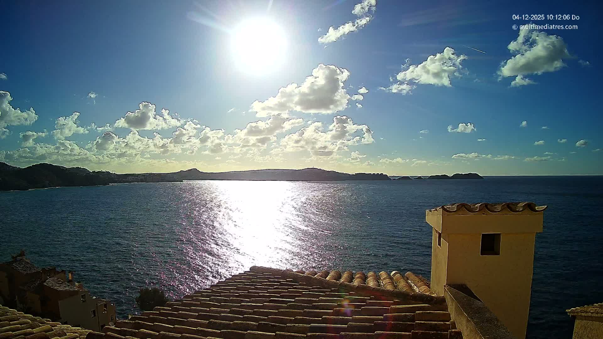 A bright, sunny coastal landscape unfolds from a high vantage point above terracotta rooftops, revealing a sparkling blue sea reflecting the sun, distant landmasses, and a clear blue sky dotted with scattered white clouds.