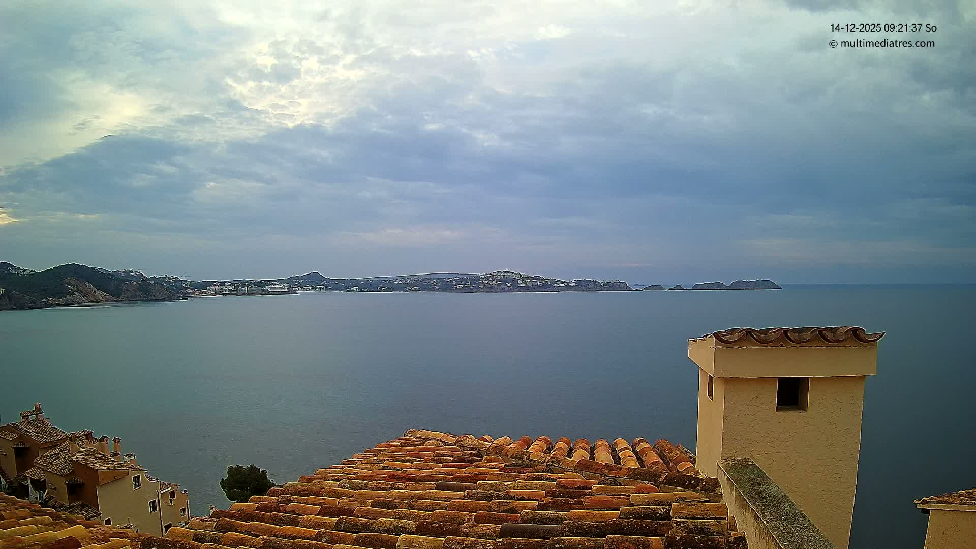 A bright, sunny coastal landscape unfolds from a high vantage point above terracotta rooftops, revealing a sparkling blue sea reflecting the sun, distant landmasses, and a clear blue sky dotted with scattered white clouds.