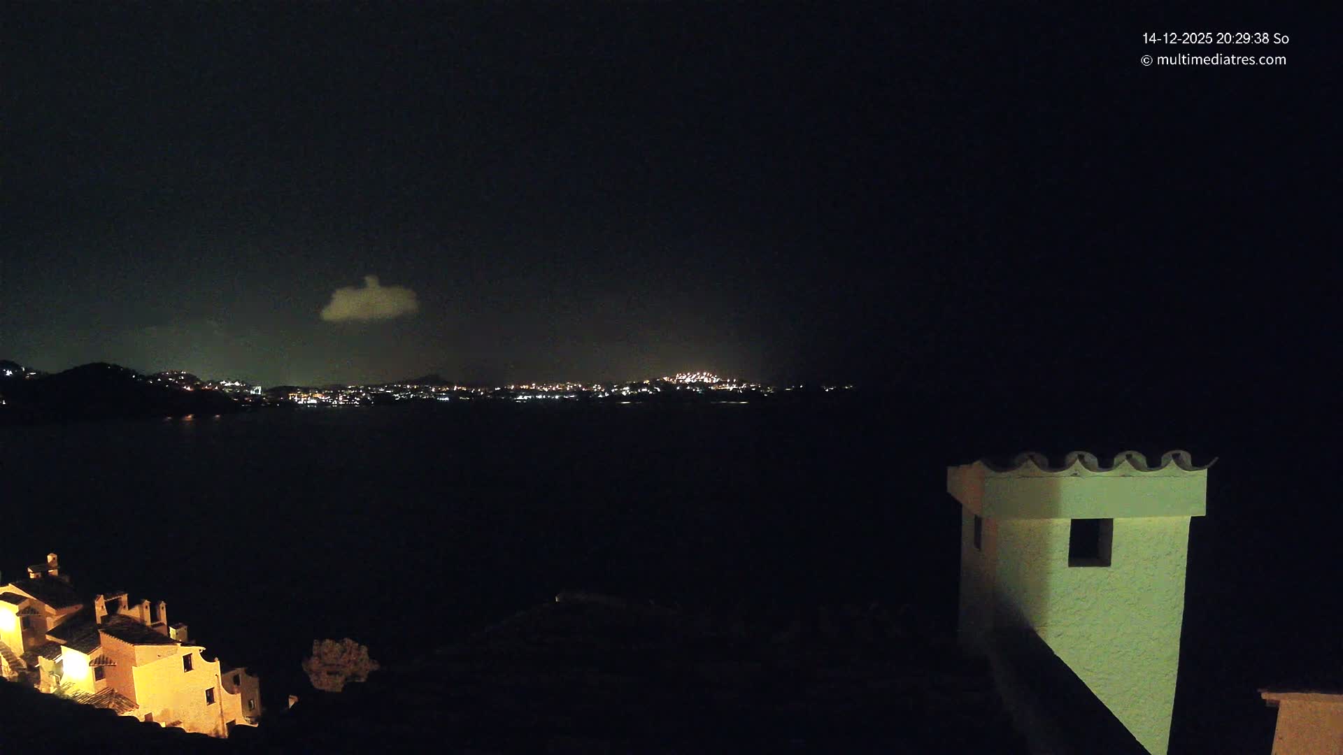 A bright, sunny coastal landscape unfolds from a high vantage point above terracotta rooftops, revealing a sparkling blue sea reflecting the sun, distant landmasses, and a clear blue sky dotted with scattered white clouds.