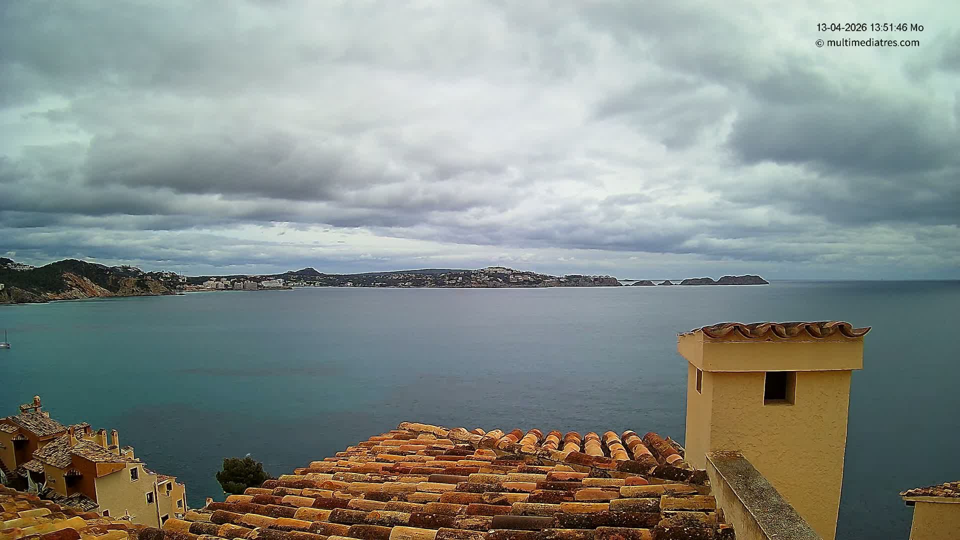 A bright, sunny coastal landscape unfolds from a high vantage point above terracotta rooftops, revealing a sparkling blue sea reflecting the sun, distant landmasses, and a clear blue sky dotted with scattered white clouds.