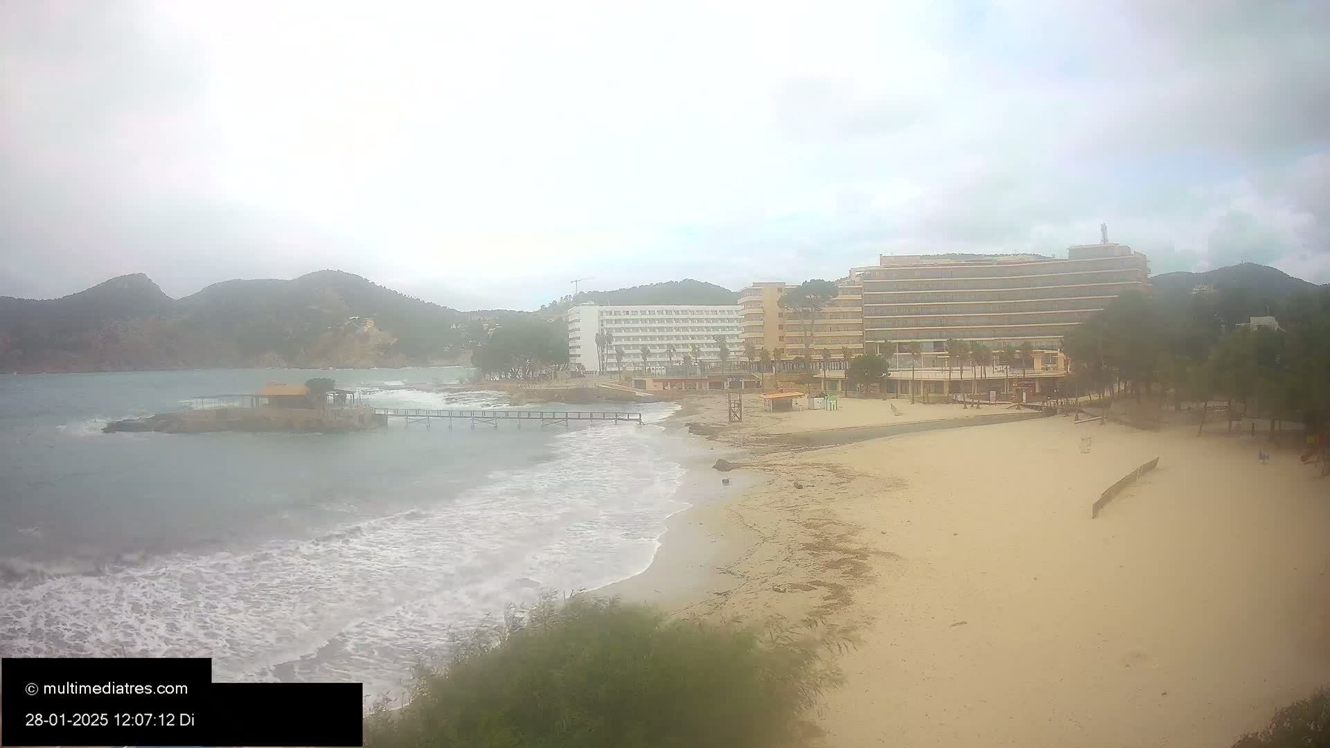 A sandy beach with choppy waves, a small pier, and several hotels is visible under an overcast sky.