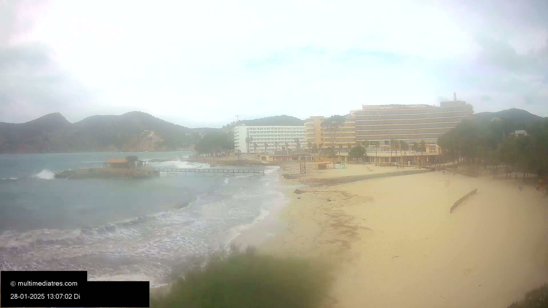 A beach with moderately sized waves crashing on the shore, in front of several hotels and a small island, under an overcast sky.