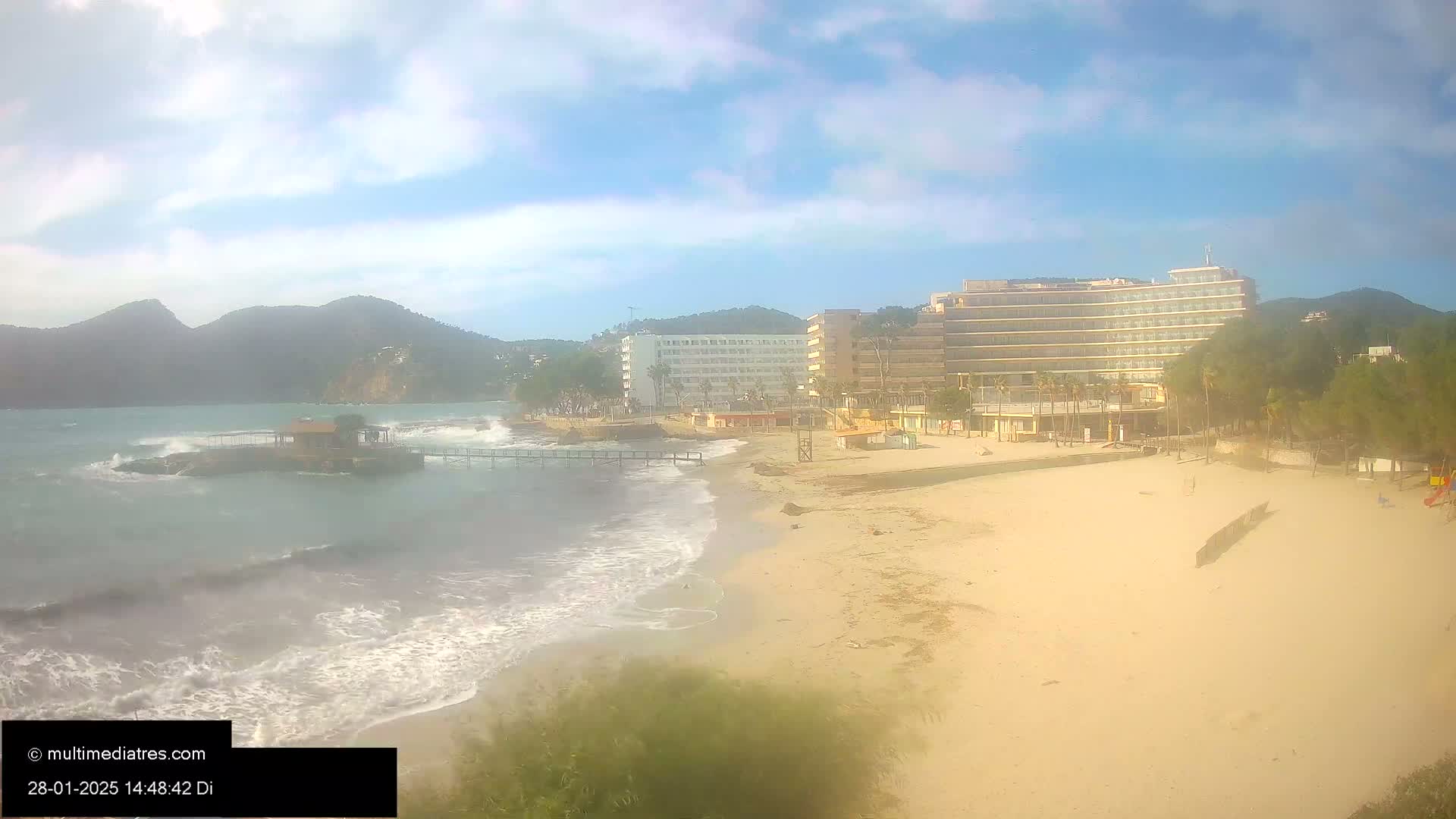 A sandy beach with moderate waves laps against the shore near a line of hotels and a small island under a partly cloudy sky.