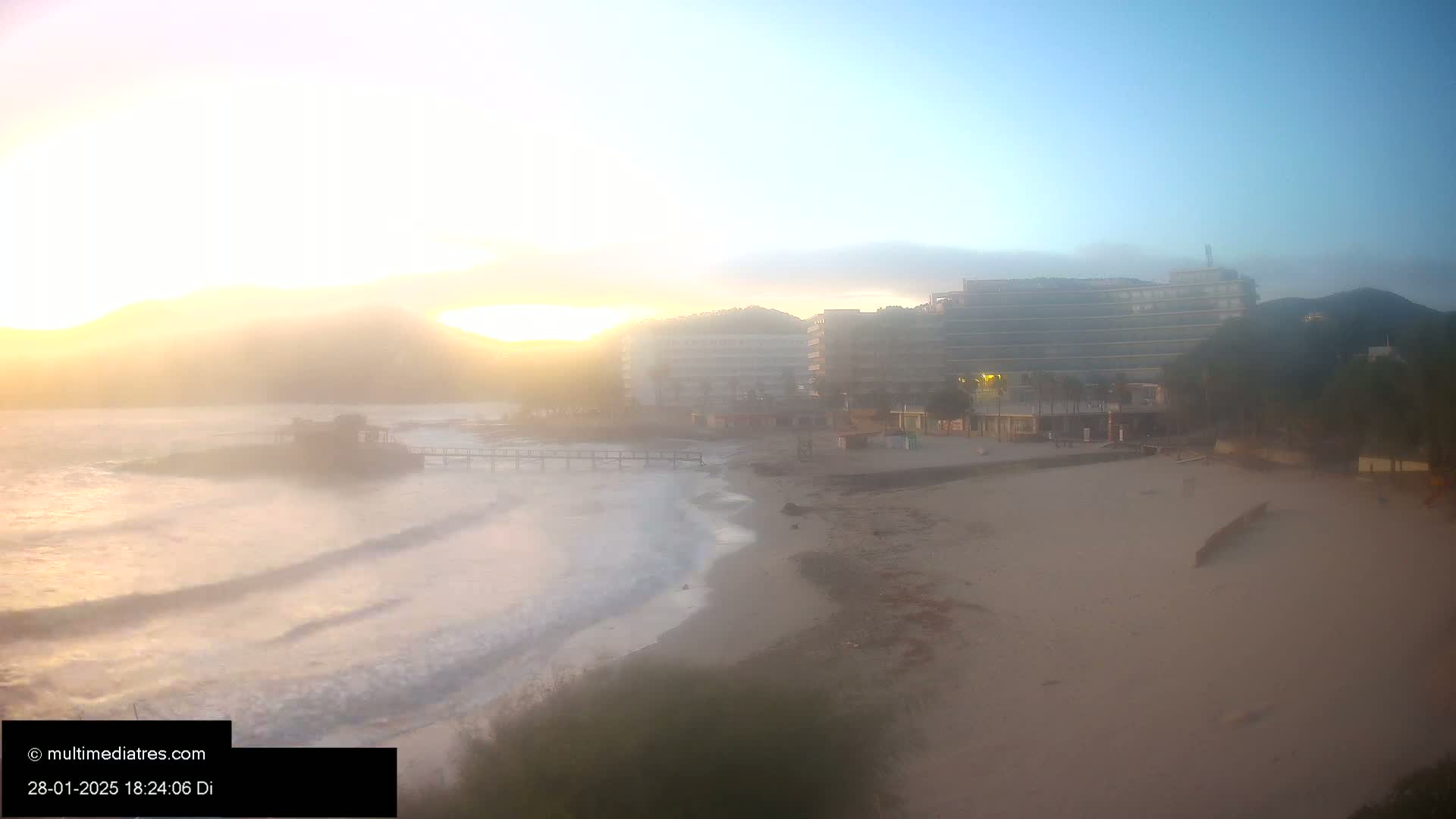 A hazy sunrise illuminates a sandy beach with gentle waves lapping the shore, featuring hotels and buildings in the background.