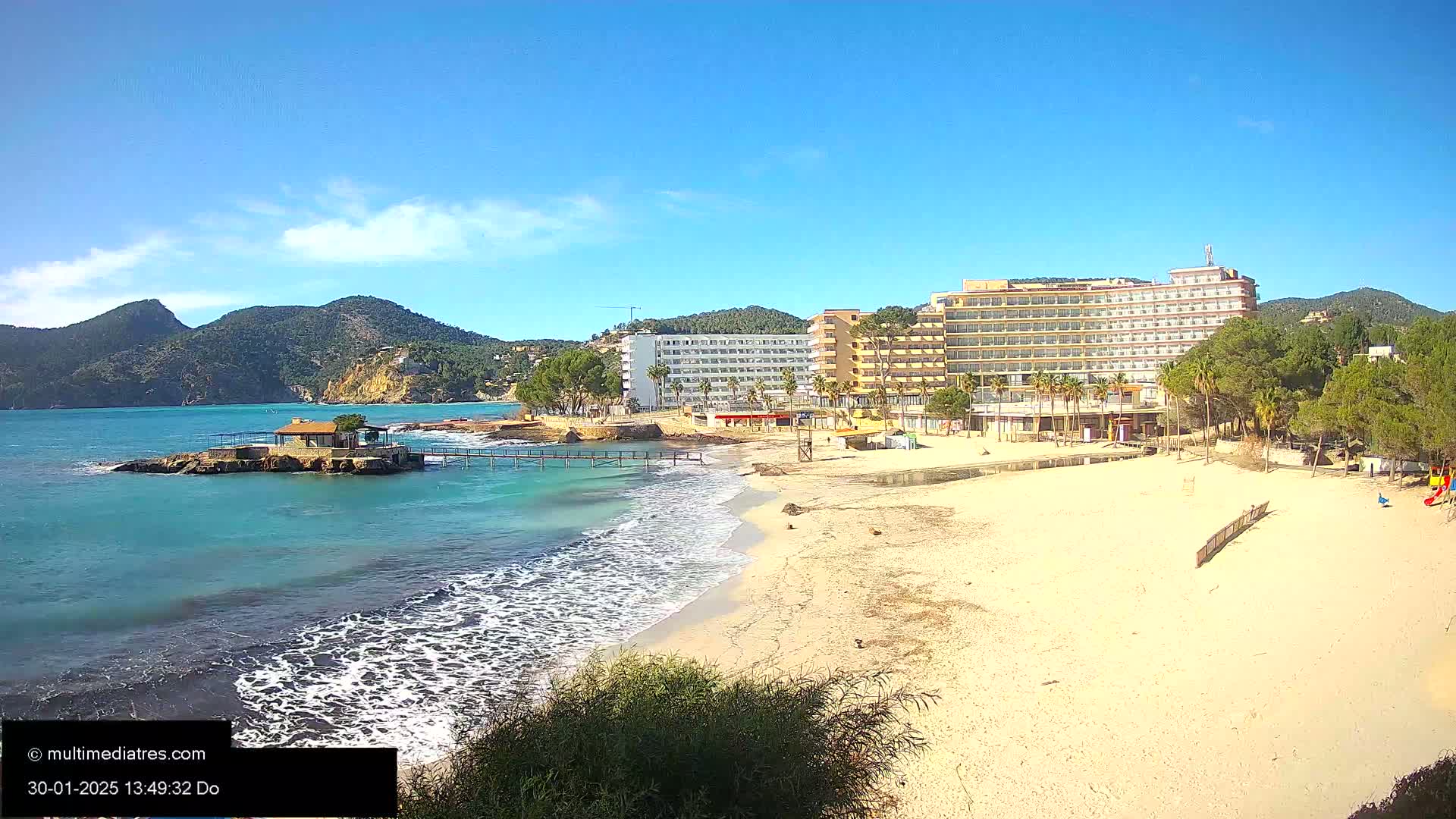 A sandy beach with turquoise water laps the shore near a row of hotels, backed by green mountains under a clear blue sky.
