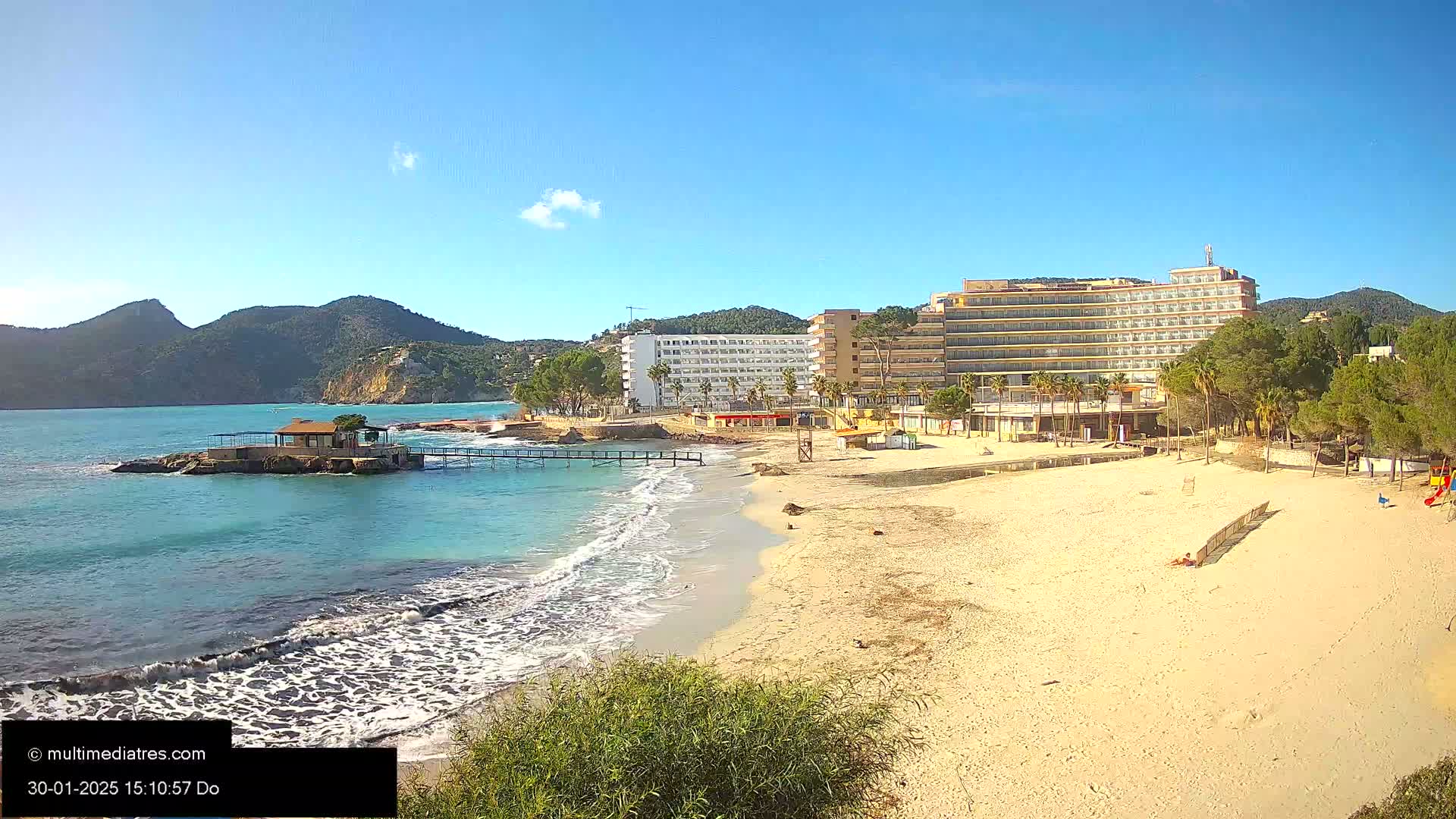 A sandy beach with gentle waves laps the shore near a hotel and small island under a clear blue sky.