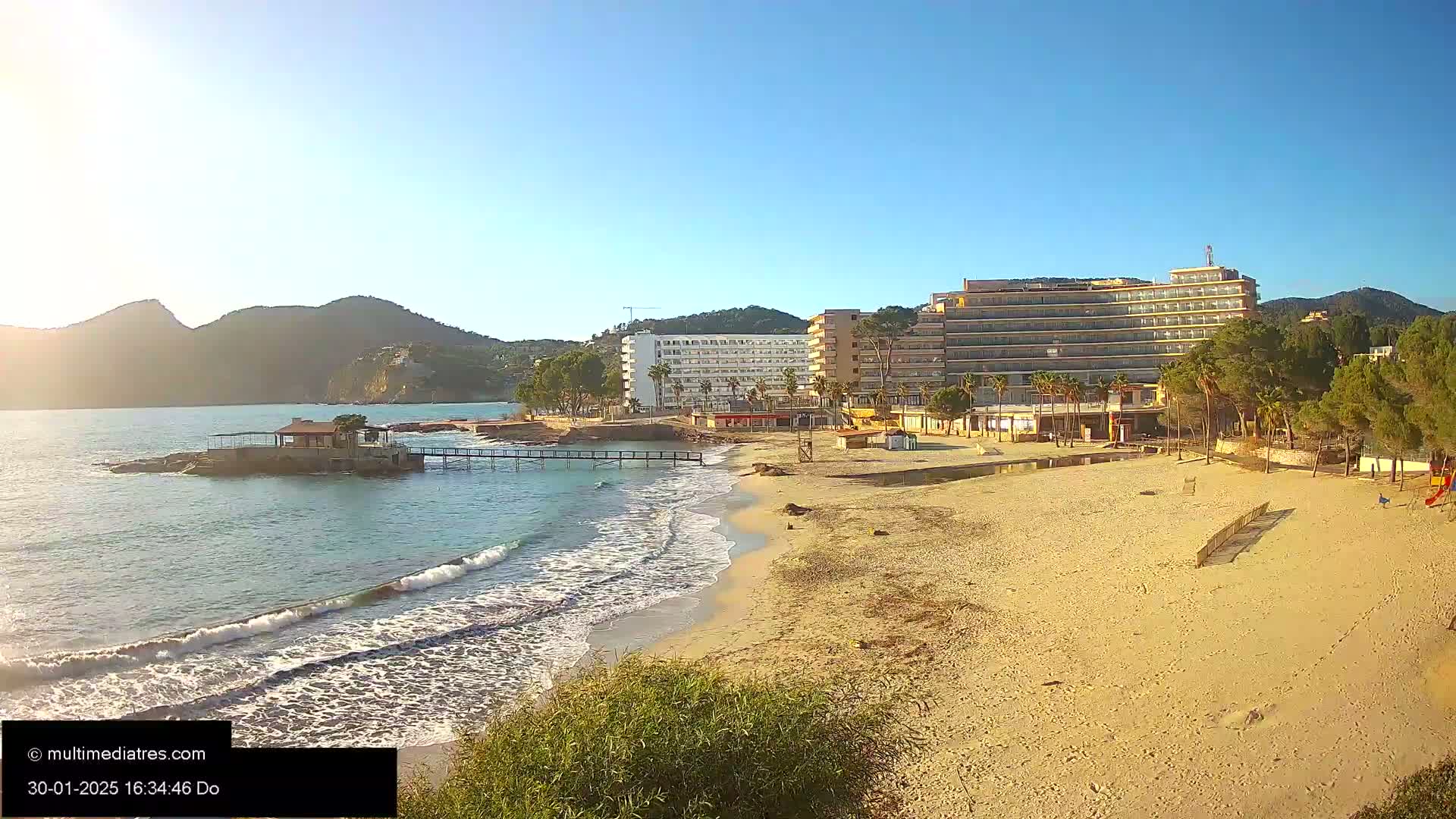 A sandy beach with gentle waves laps the shore near several hotels and a small building on a rocky outcrop, all under a clear, sunny sky, backed by mountains.