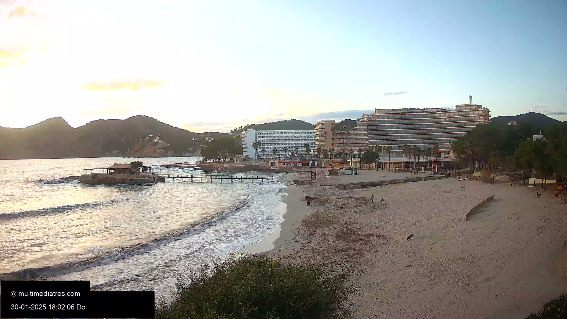 A sandy beach with gentle waves laps at the shore near several hotels and a small structure on the water under a partly cloudy, pale sky.