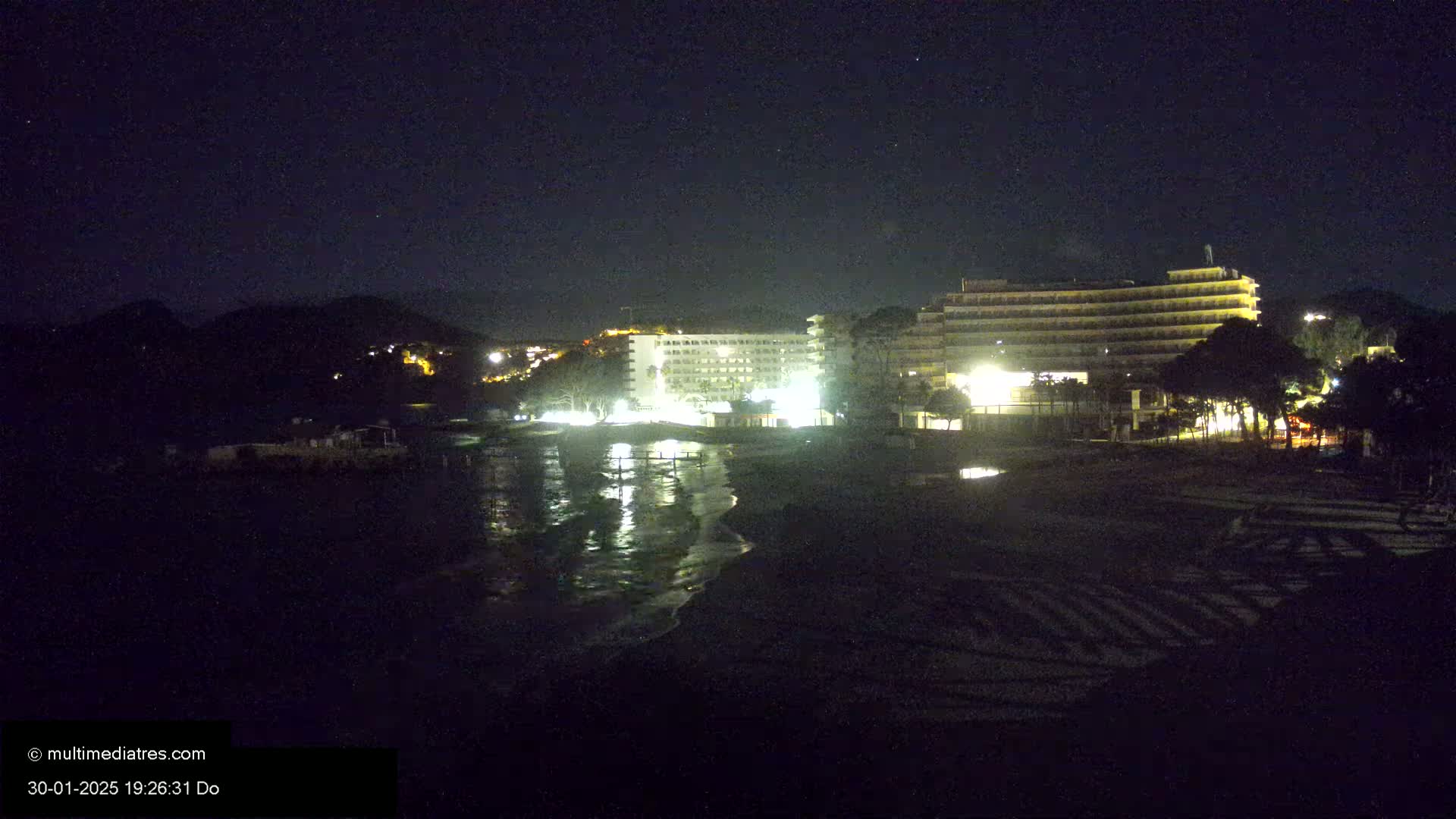 At night, a dark beach with lit hotels and a calm, reflecting body of water sits before a hill under a clear, starry sky.
