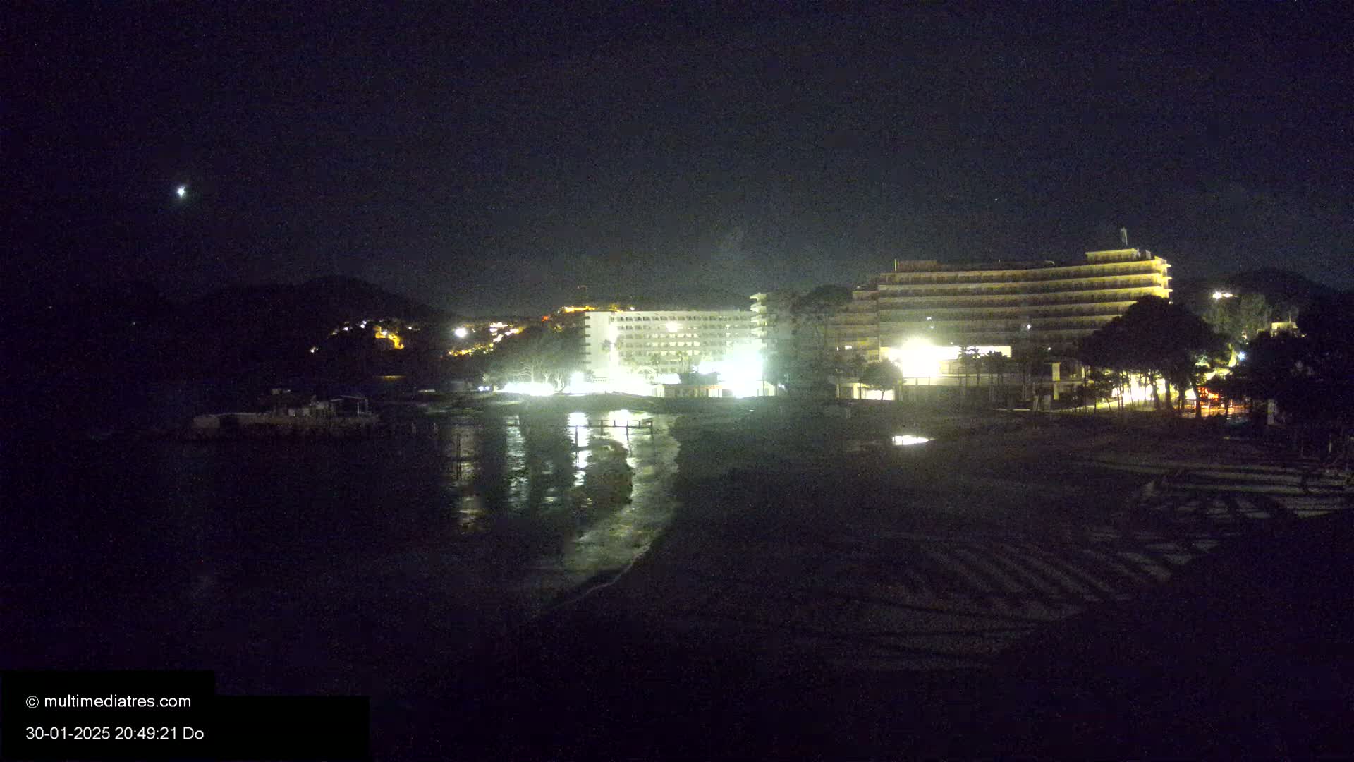 A nighttime view of illuminated hotels and a beach on a calm sea under a clear, dark sky.