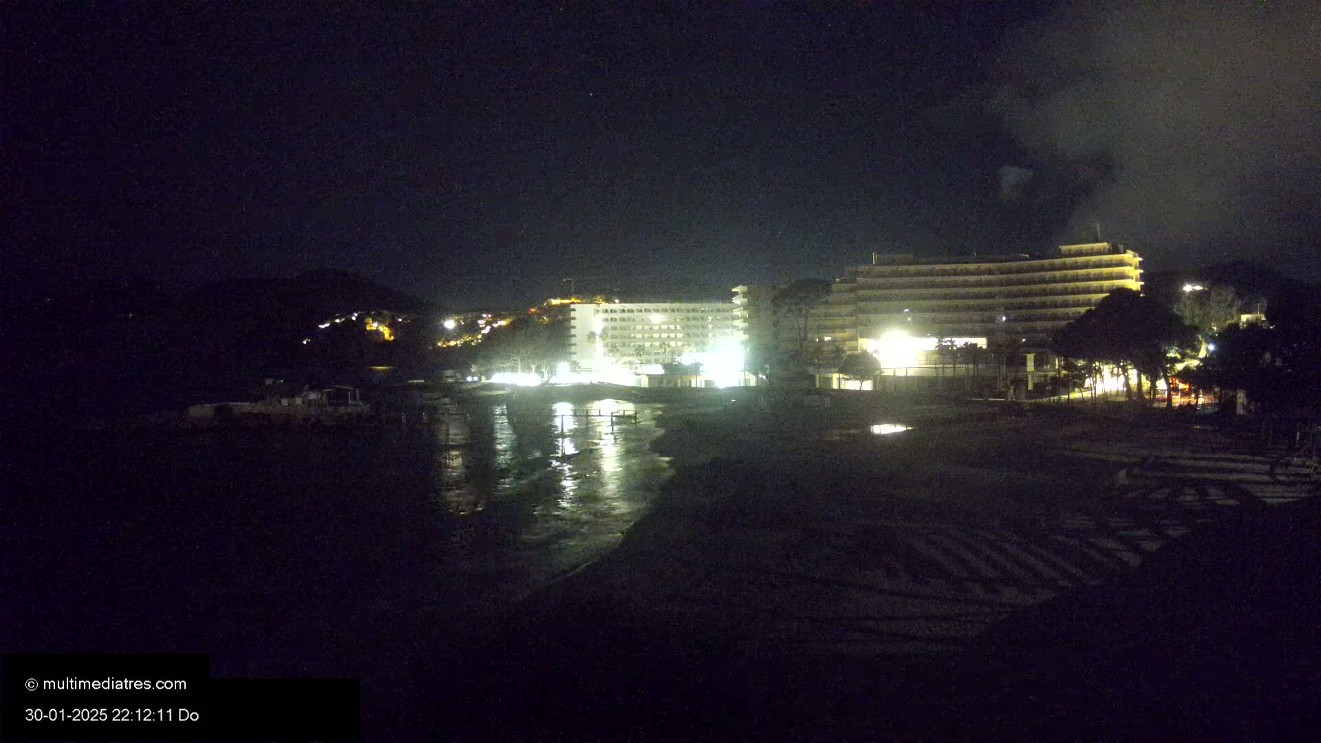 At night, illuminated hotels are situated near a dark beach with calm water reflecting the lights.