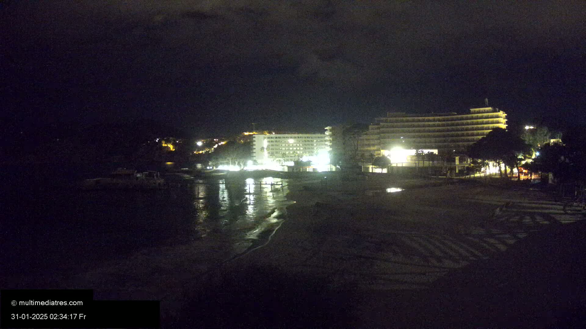 At night, two illuminated hotels are situated near a dark, sandy beach and calm, reflective water.