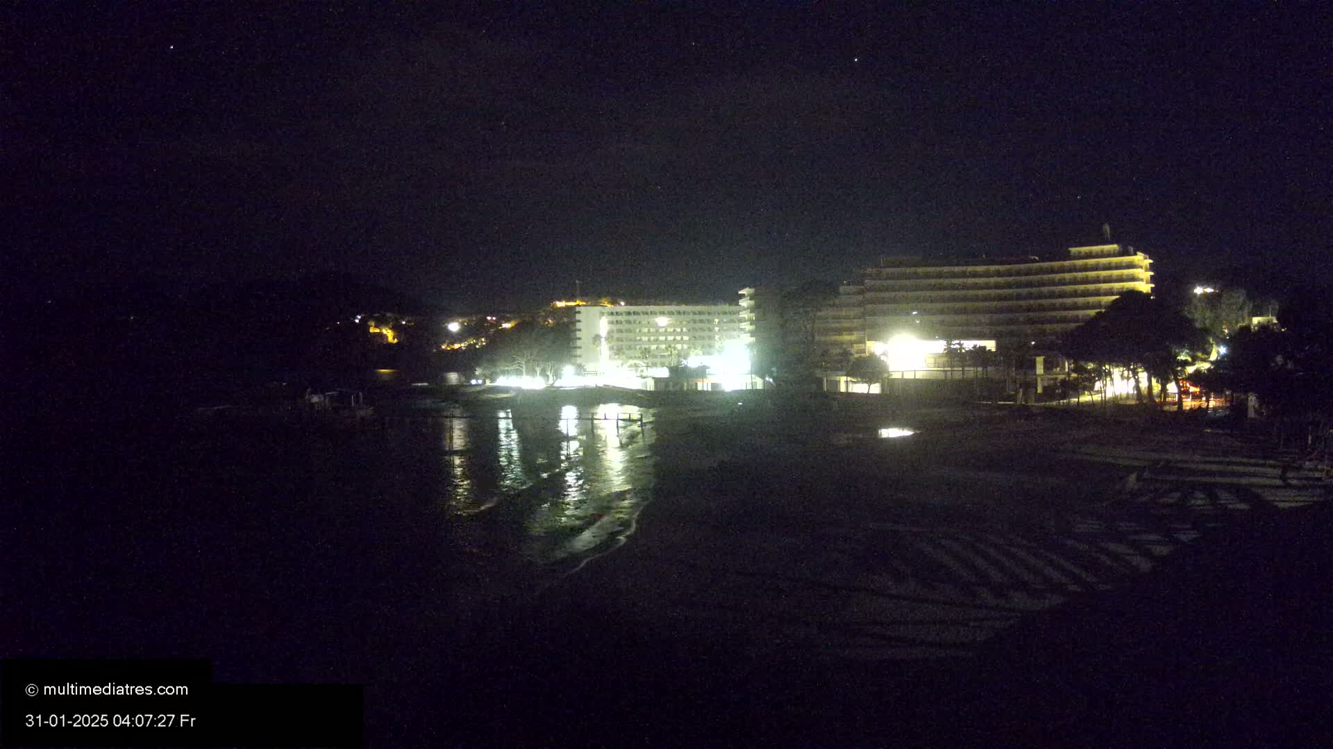 Two brightly lit hotels are situated on a dark, calm beach at night under a clear, starry sky.