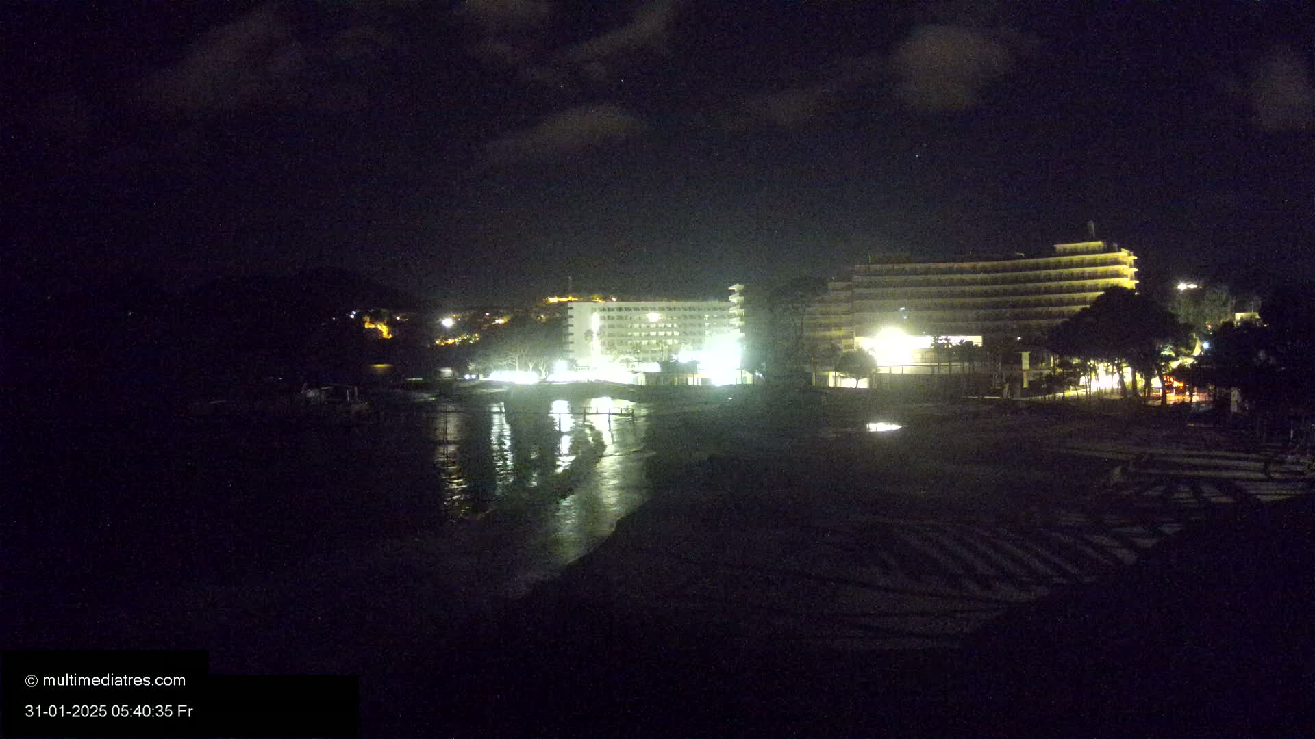 A nighttime view of a beach with illuminated hotels and calm water reflecting lights.