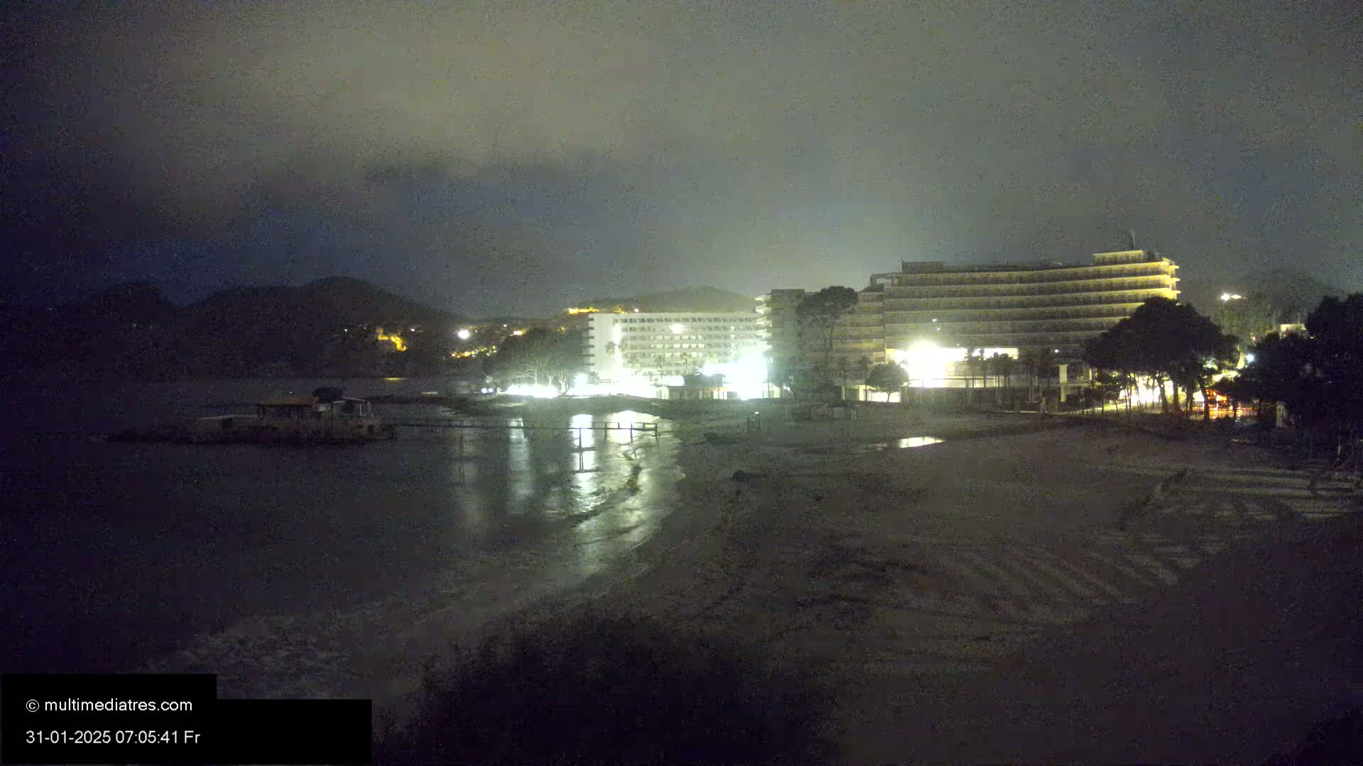 At night, under a cloudy sky, two illuminated hotels sit on a beach next to calm water.