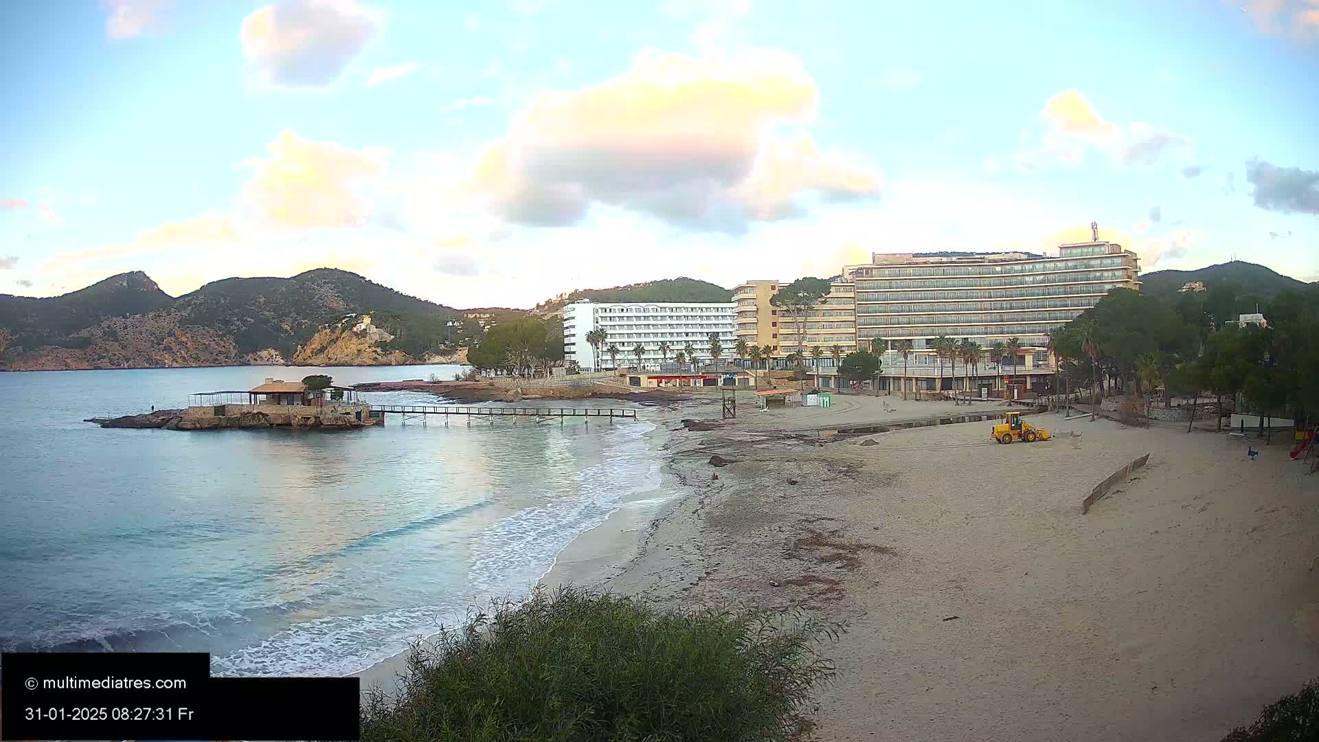A sandy beach with calm waves laps the shore, near a small island with a building and several large hotels under a partly cloudy sky.