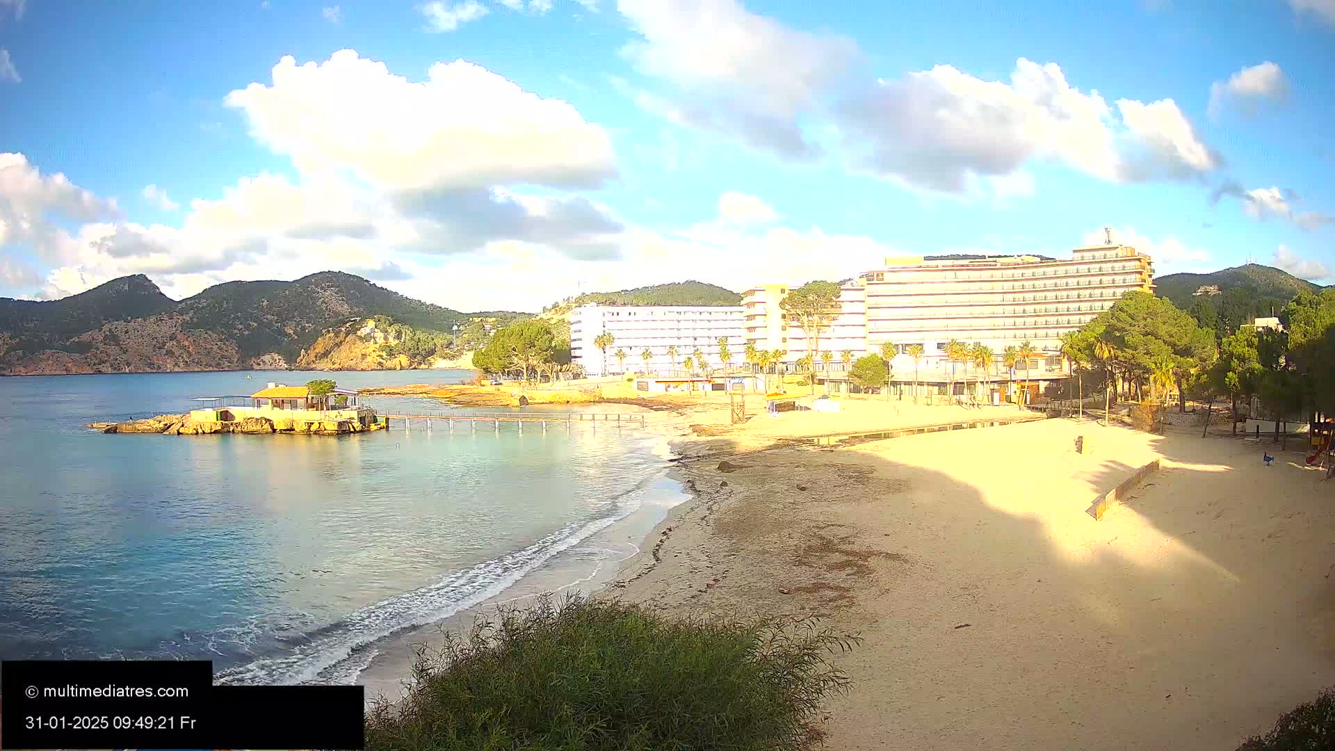 A sandy beach with calm, clear water laps the shore near a large hotel, under a partly cloudy, sunny sky, with mountains visible in the distance.