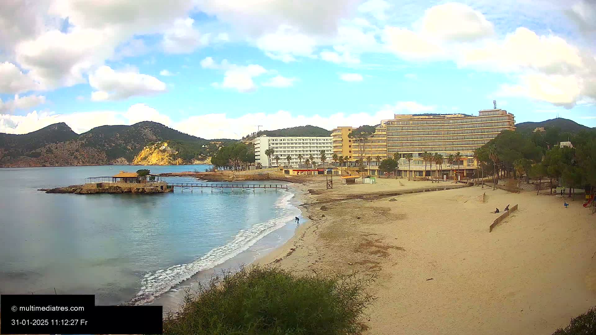 A sandy beach with a few people on it stretches out in front of several hotels and a small island with a building on it, all under a partly cloudy sky.