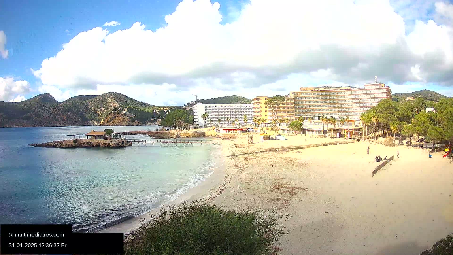 A sandy beach with a couple of hotels and a small island with a building on it is visible under a partly cloudy sky.
