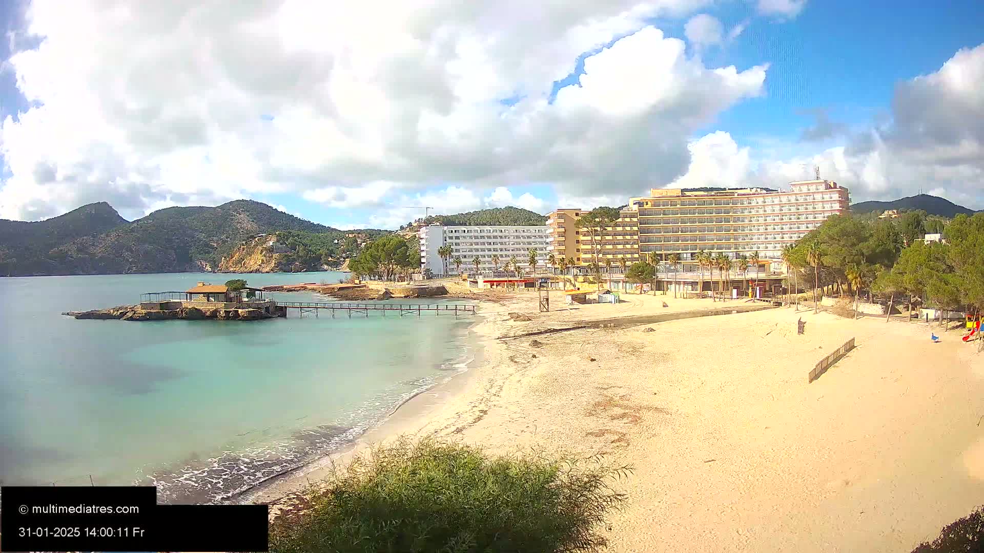 A sandy beach with turquoise water stretches out to a calm bay, backed by hotels and lush green hills under a partly cloudy sky.