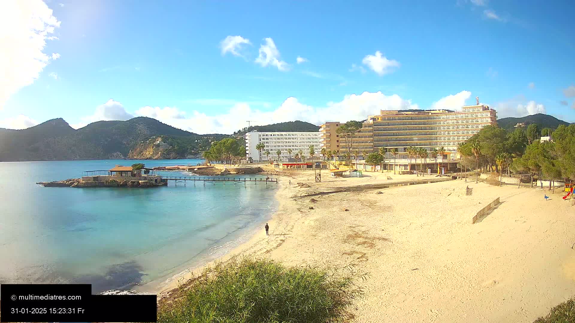 A sandy beach with a large hotel and a small island with a pavilion in the background, under a mostly sunny sky with some clouds.