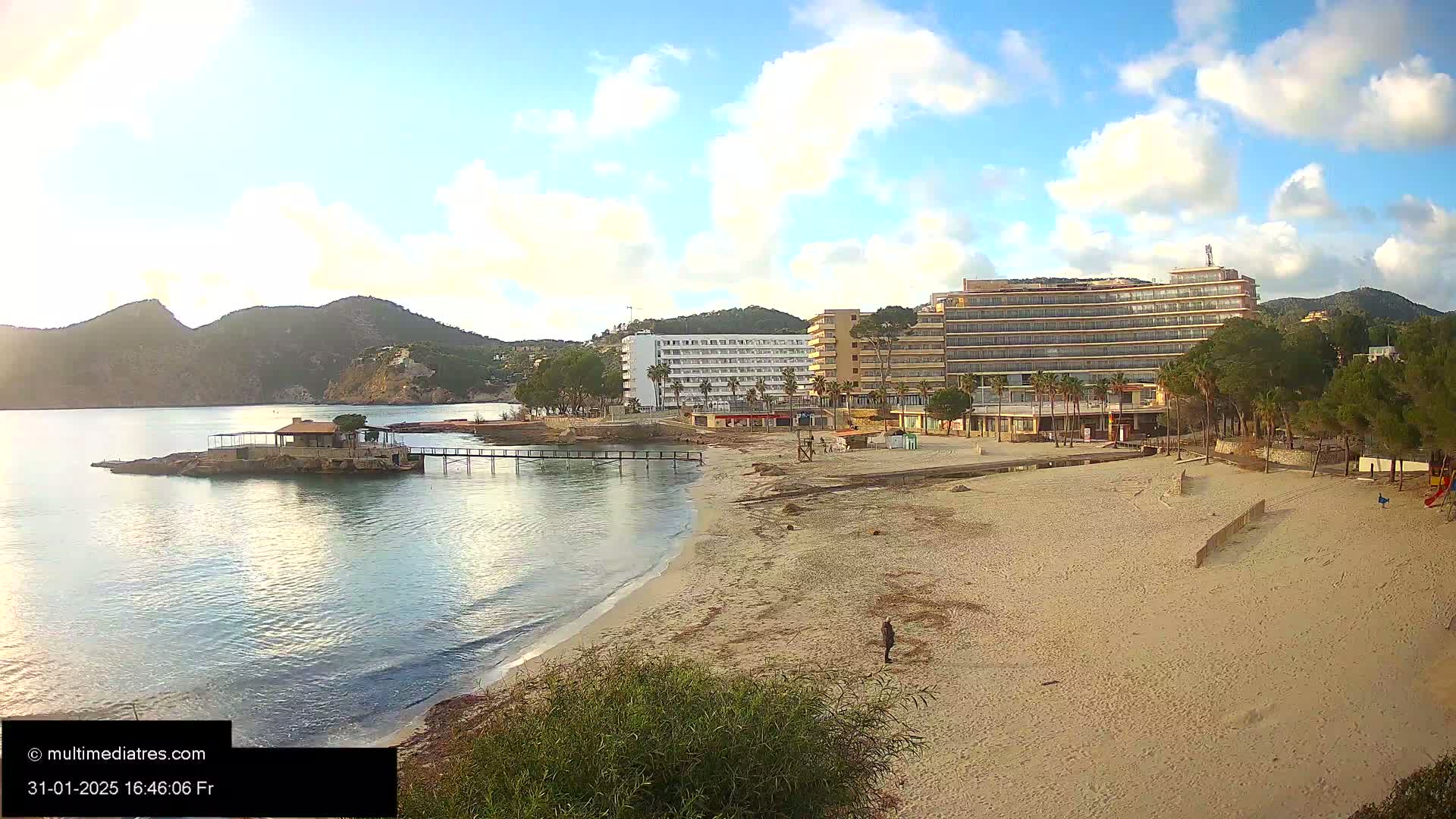 A sandy beach with calm water, a small pier leading to a building on a rocky outcrop, and large hotels behind it under a partly cloudy sky.