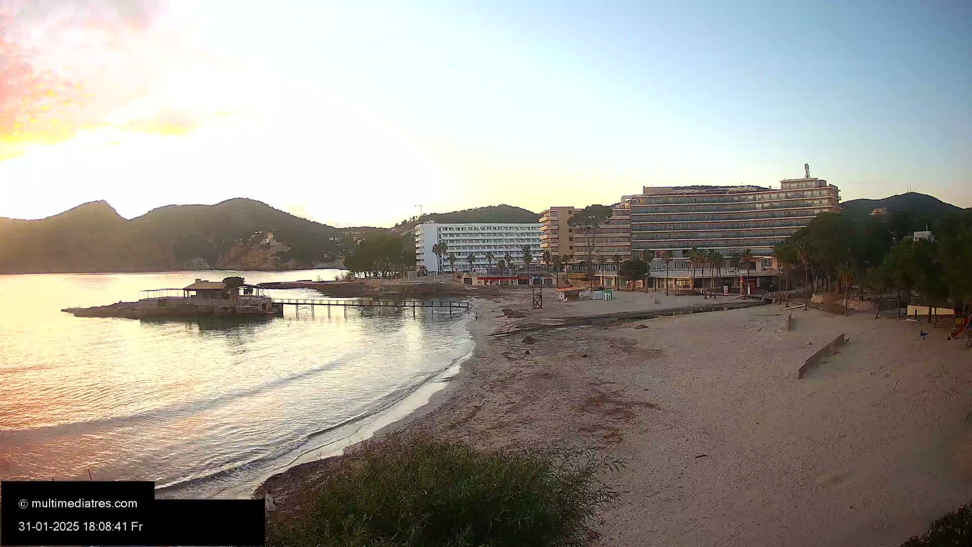 A sandy beach at sunset, with calm ocean waves lapping the shore, lies in front of several hotels and a small structure on a rocky outcropping, all nestled against a backdrop of hills.