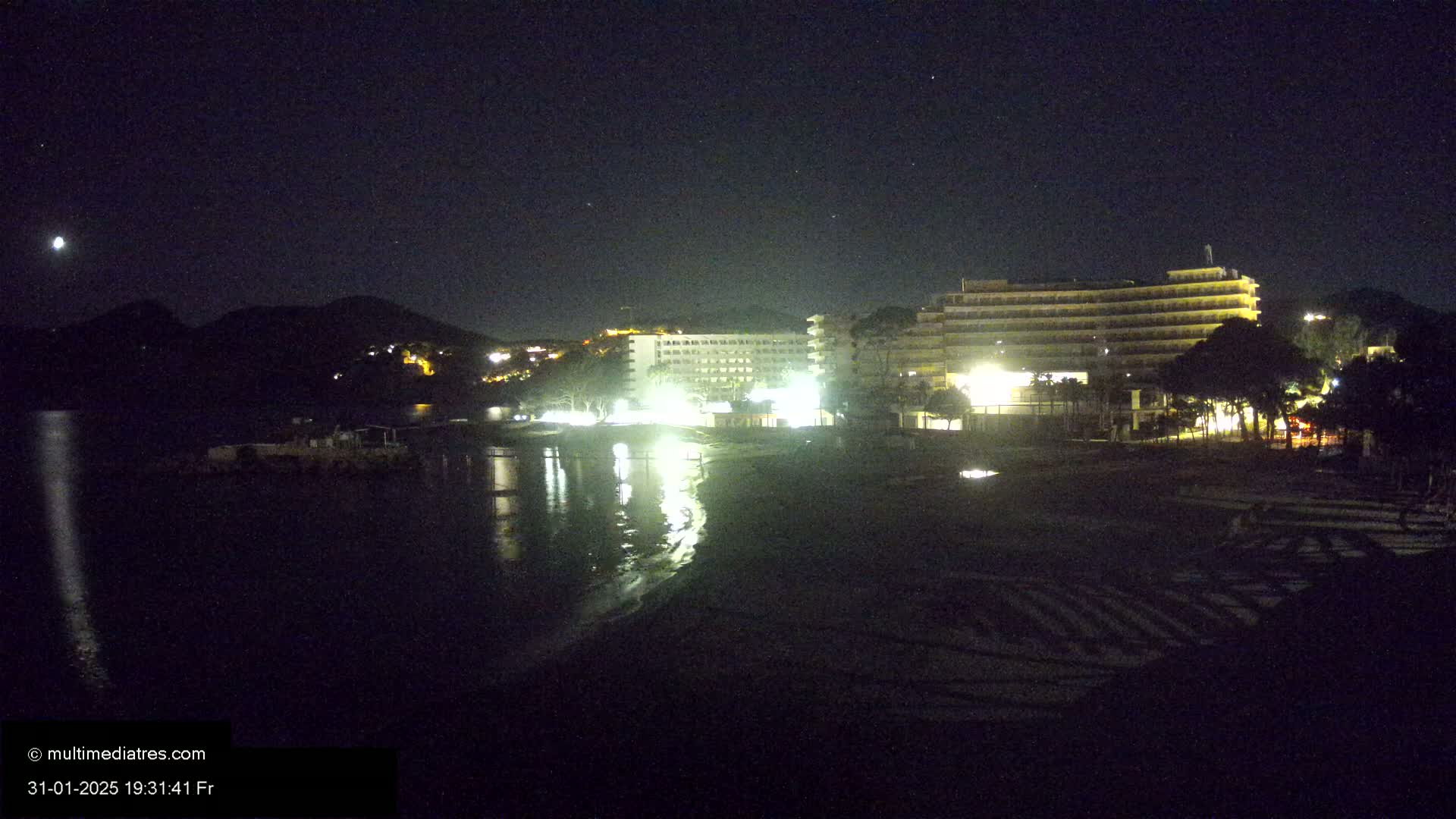 A nighttime view of a beach with illuminated hotels and calm water under a clear, starry sky.