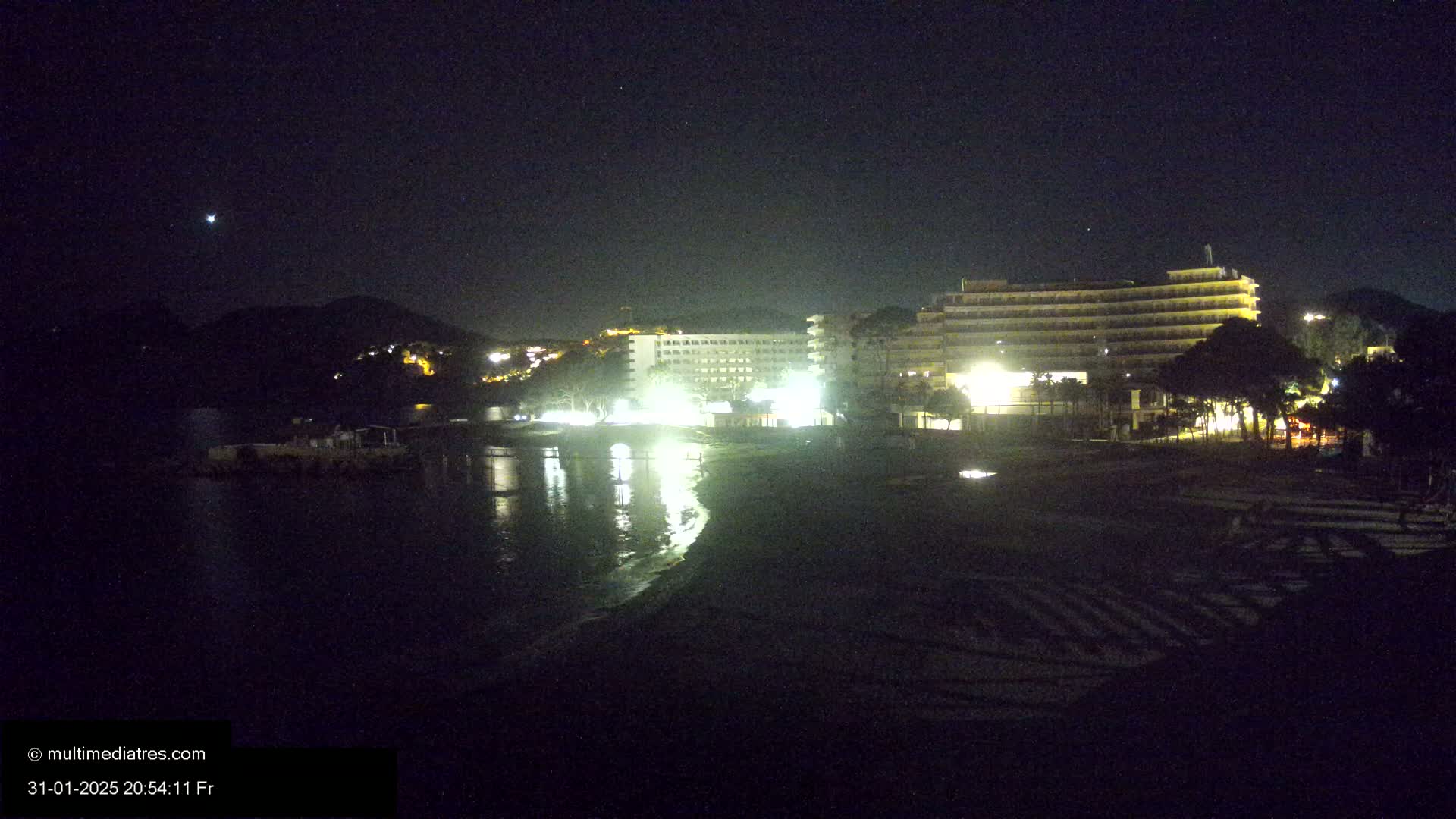 A nighttime view of a beach with illuminated hotels and calm water under a clear, starry sky.