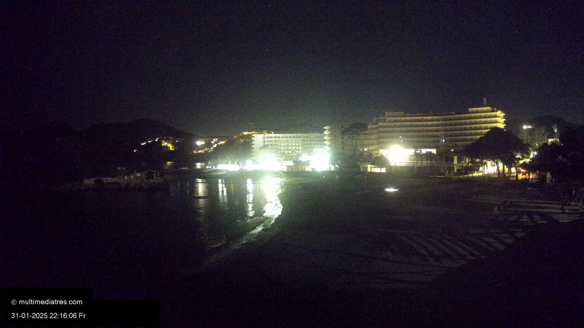 A nighttime view of a beach with brightly lit hotels and calm water reflecting the lights.