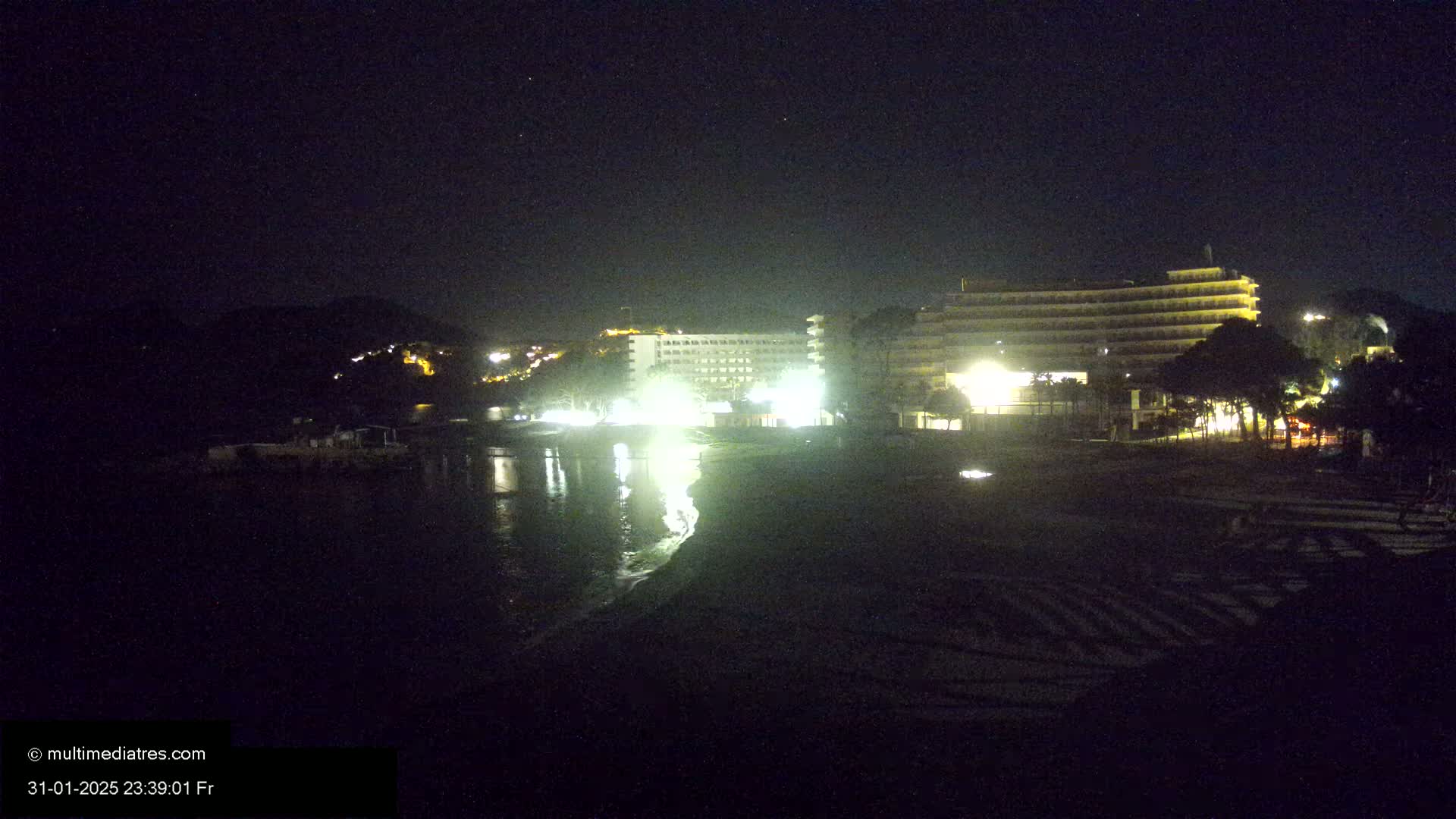 At night, brightly lit hotels stand on a dark beach next to a calm body of water with hills visible in the background.
