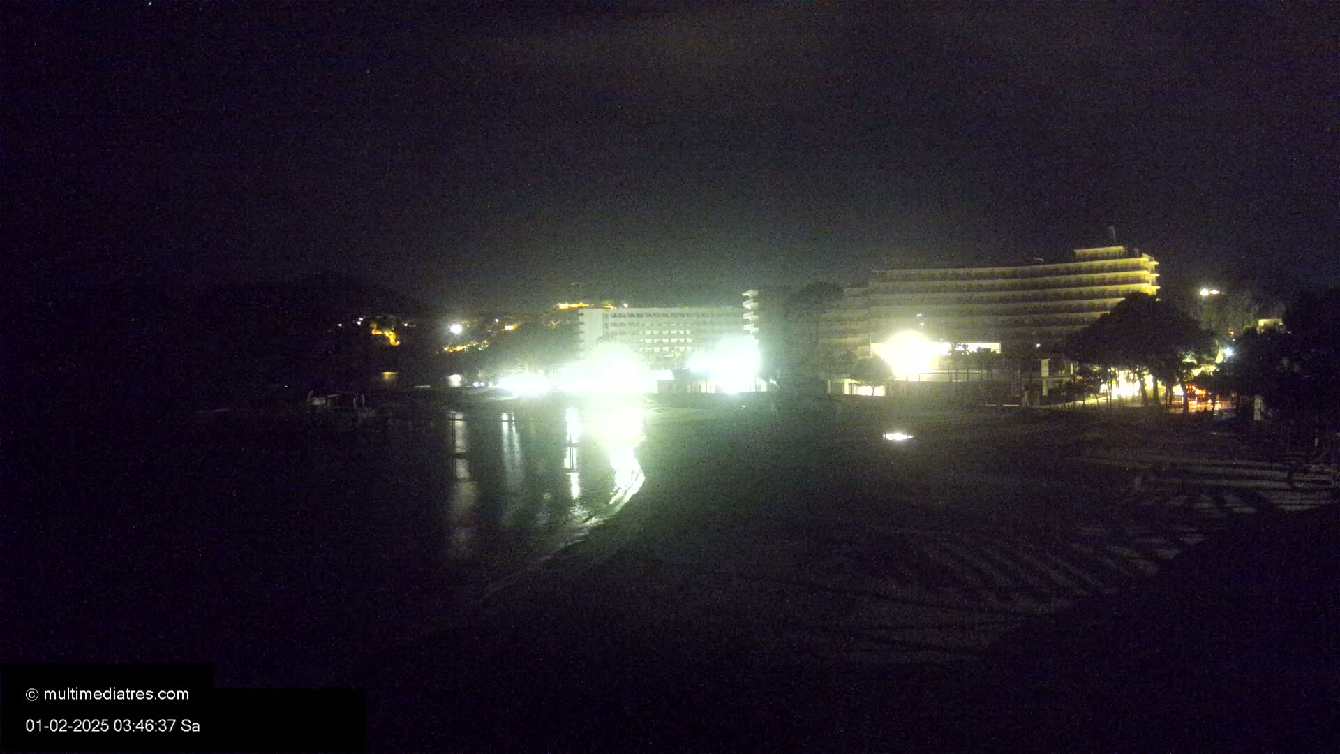 A nighttime view of a beach with brightly lit hotels and calm water reflecting the lights.