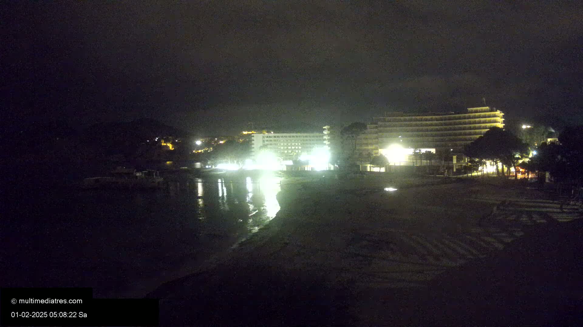 A nighttime view of a beach with illuminated hotels and calm water reflecting the lights.