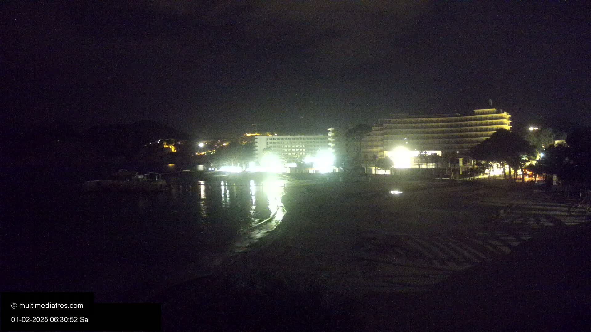 At night, a dark beach with two brightly lit hotels and a calm body of water is visible under a clear, dark sky.