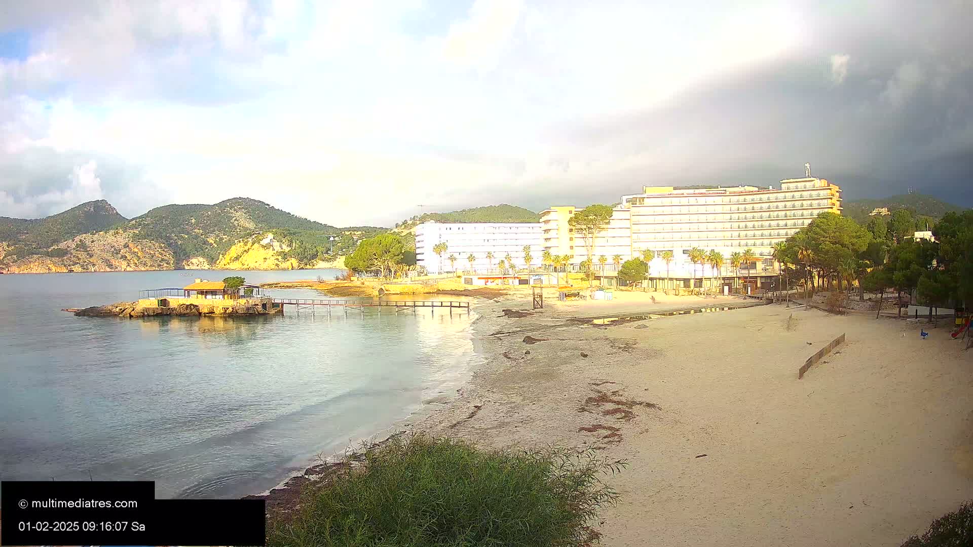 A sandy beach with a large hotel and calm, clear water is visible under a partly cloudy sky.