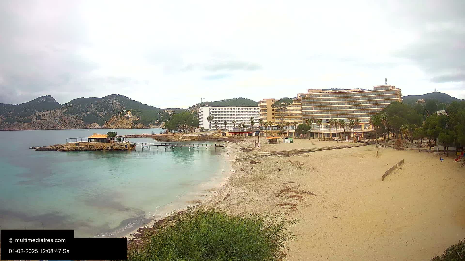 A sandy beach with calm, light-blue water stretches toward two large hotels situated near rocky hills under an overcast sky.