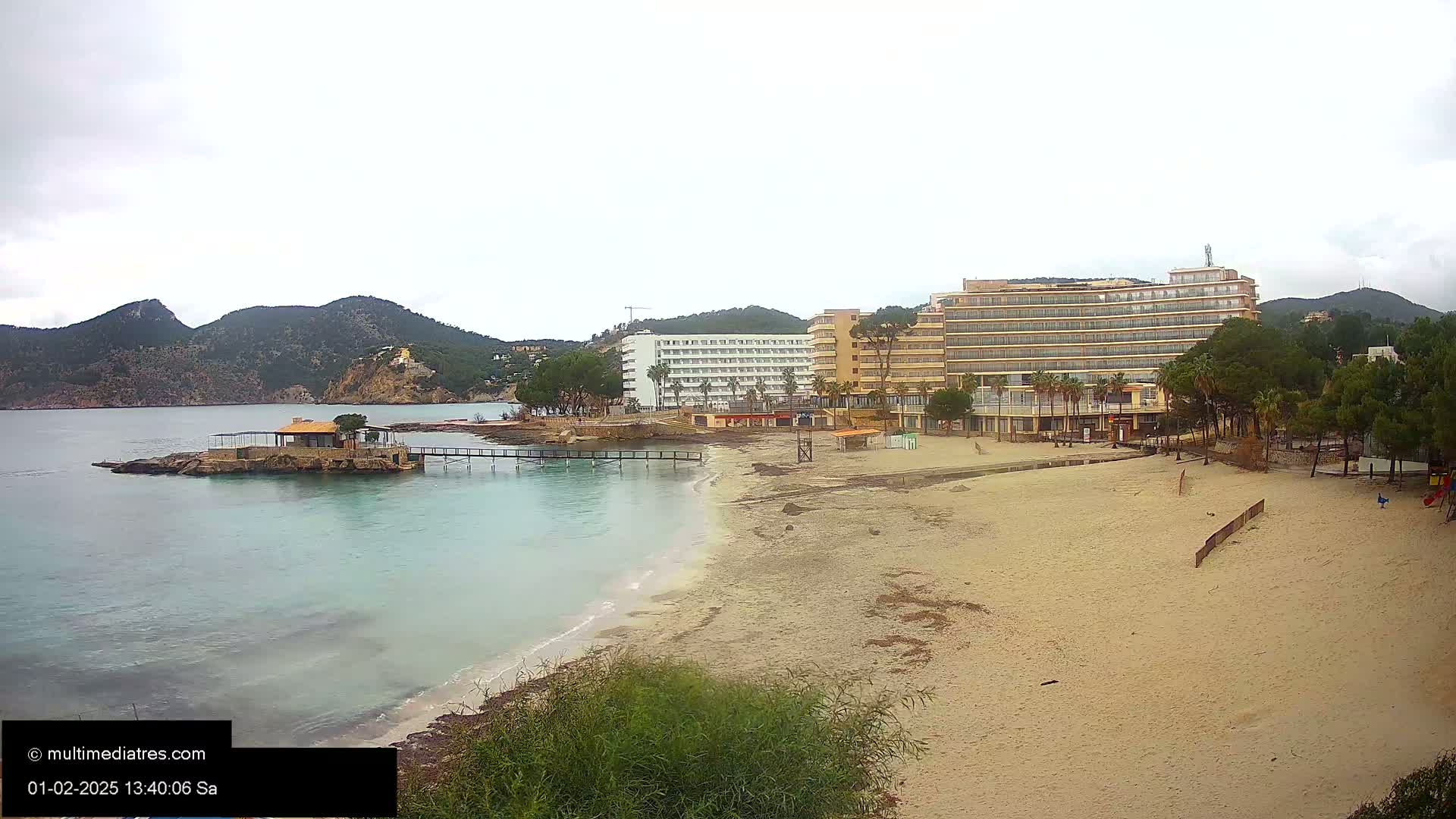 A sandy beach with calm, light-blue water stretches towards two large hotels under an overcast sky, backed by low, green mountains.