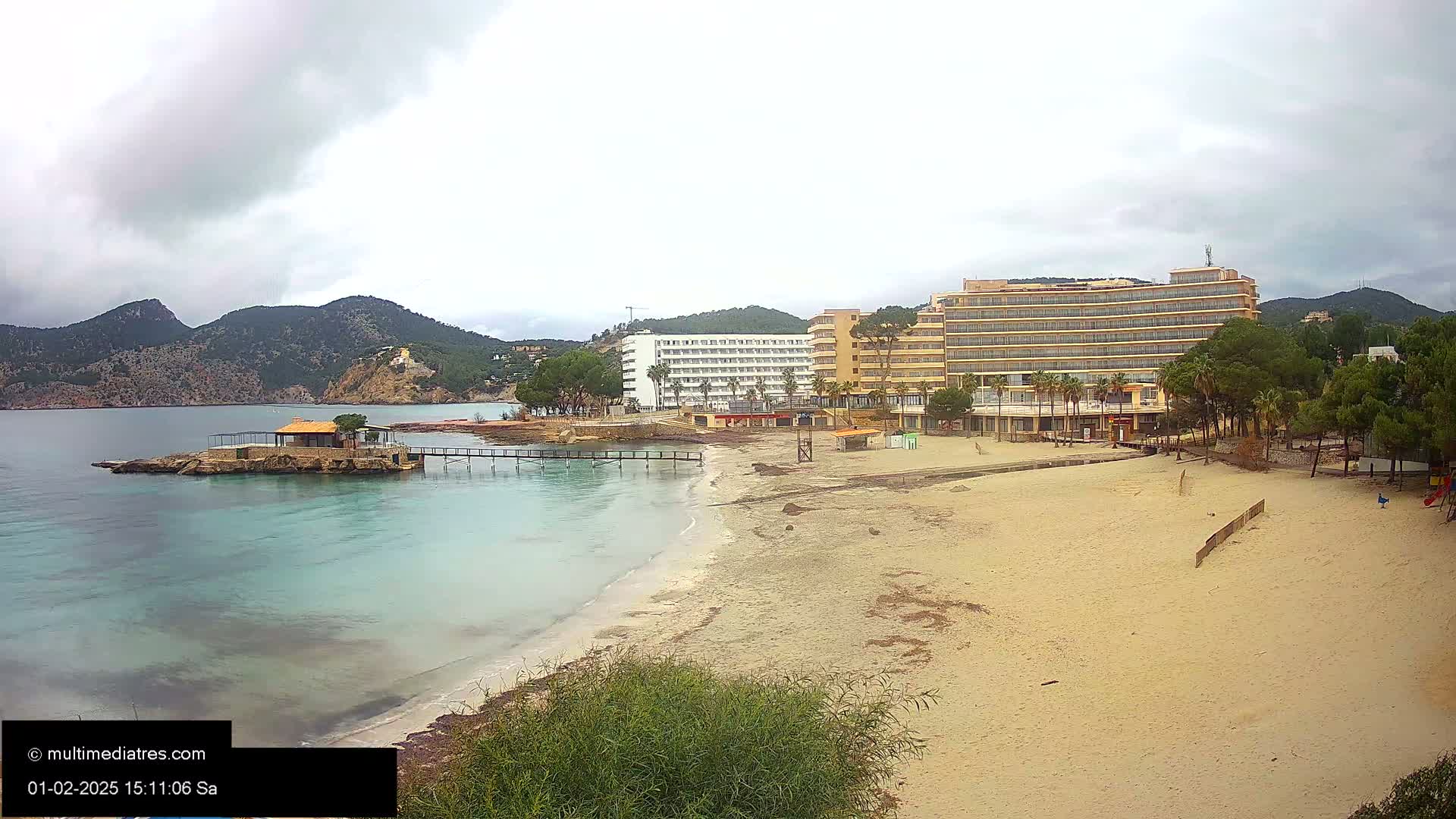A sandy beach with two hotels and a small hut on a pier, situated beneath overcast skies and mountains in the background.