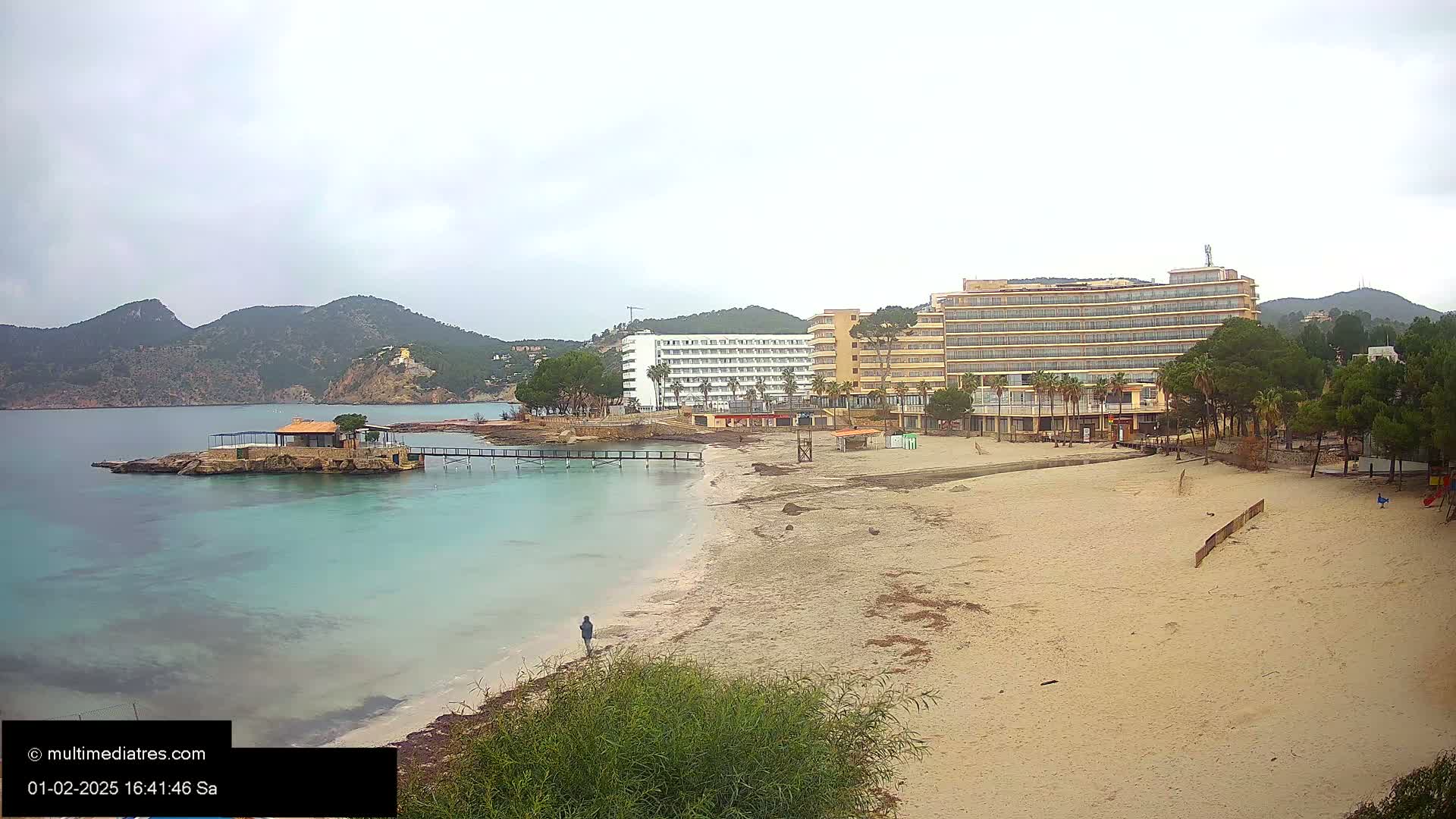 A sandy beach with a few people and several hotels behind it, all under an overcast sky, is situated next to calm, turquoise water and mountains in the background.