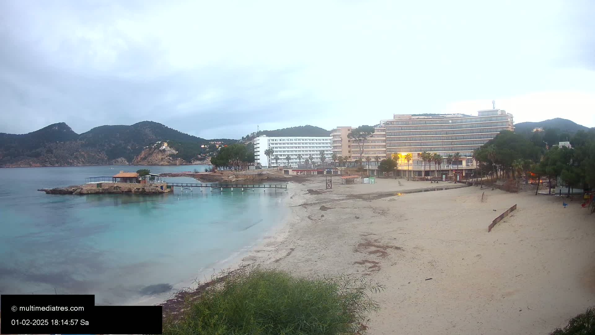 A sandy beach with calm, turquoise water, two hotels, and a small island with a building on it under an overcast sky.